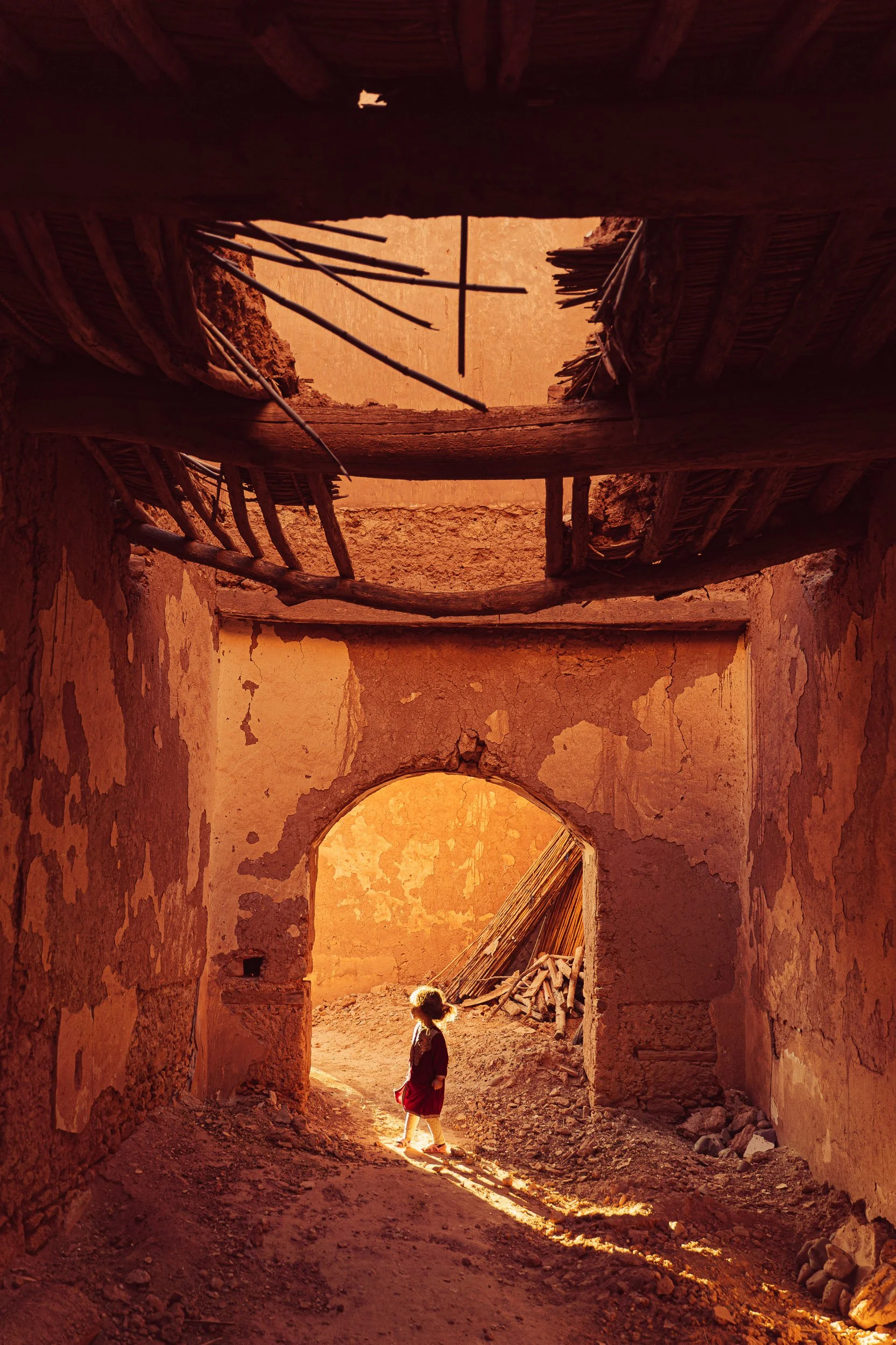 A young girl in a red skirt and dark top walking through the ruins of an old, dilapidated building with walls and roof made of mud and sticks, illuminated by warm sunlight.