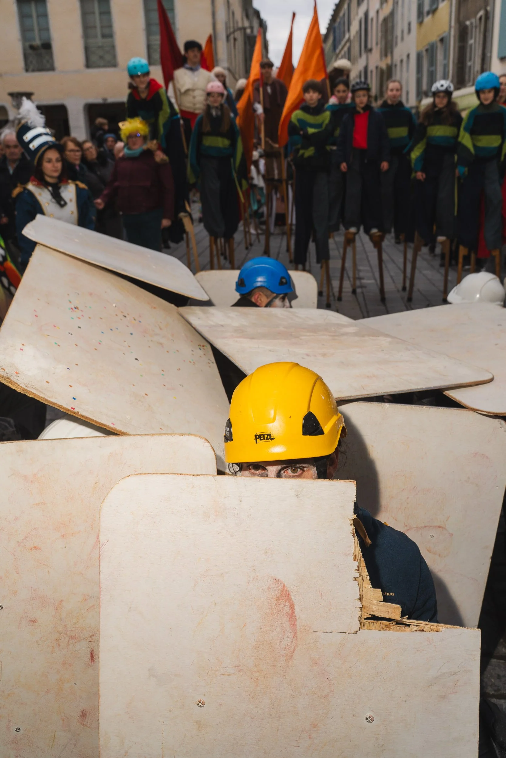 People in a street, wearing helmets, behind a makeshift wooden barrier, with others on stilts in the background holding flags.