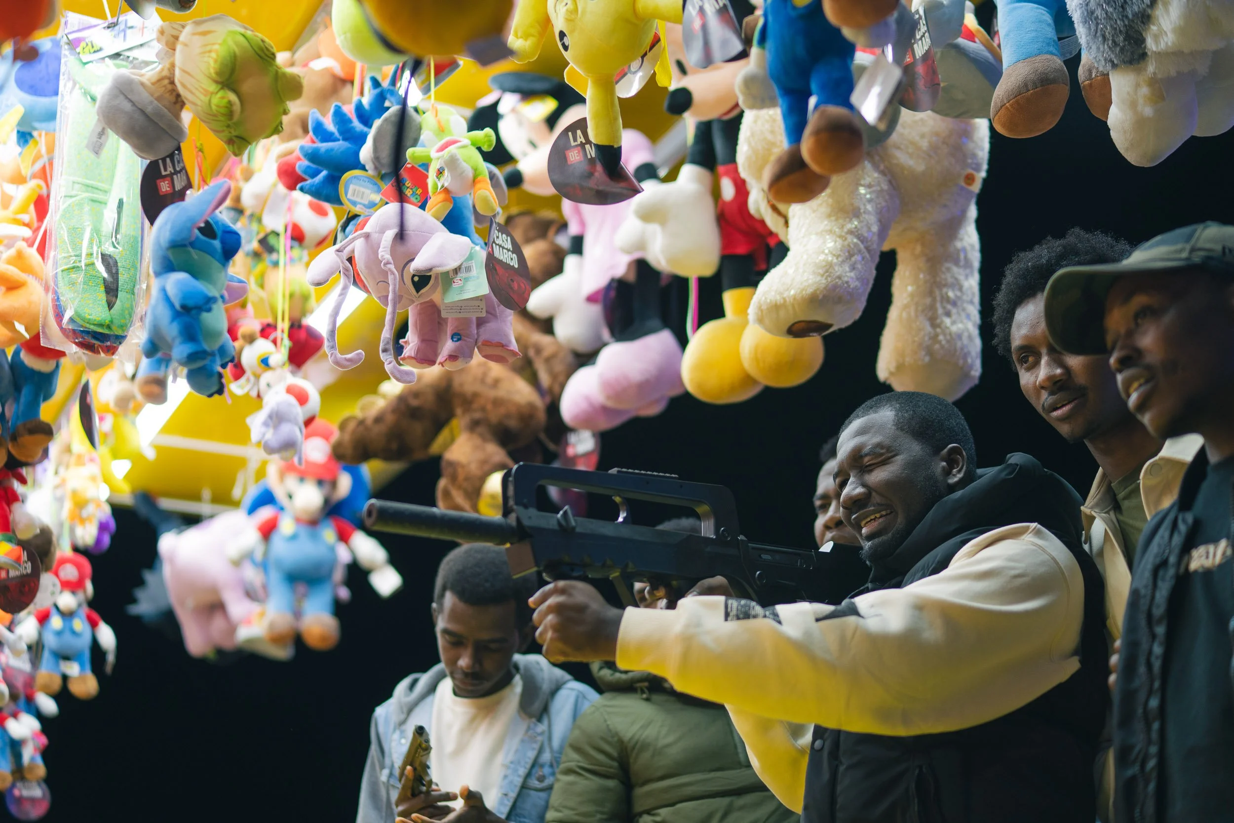 People playing a whack-a-mole game with plush toys at an arcade or fair, with some individuals appearing to aim a toy gun at the stuffed animals hanging from above.