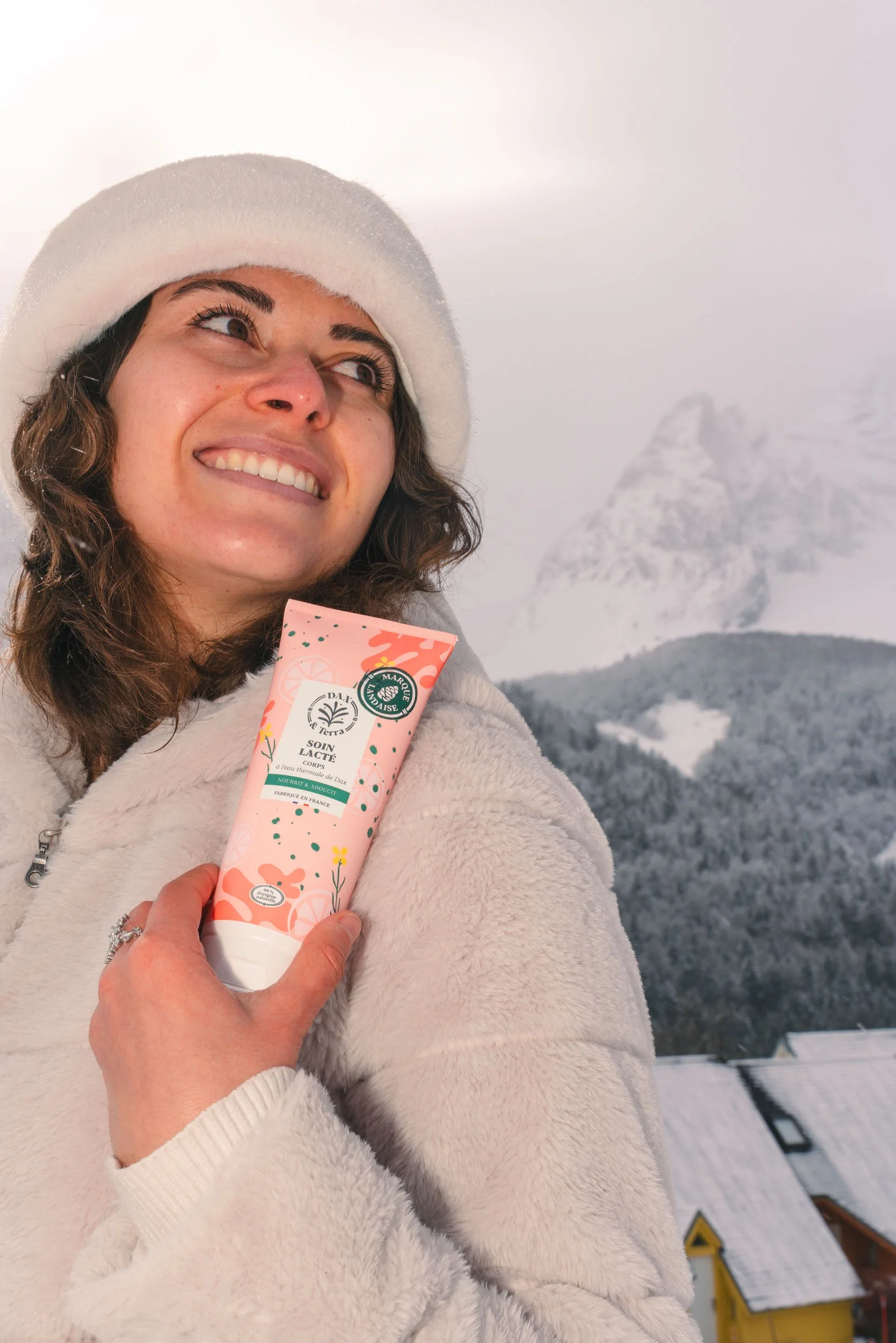 A woman wearing a white fuzzy coat and a white hat, smiling and holding a pink skincare product in front of snow-covered mountains and trees.