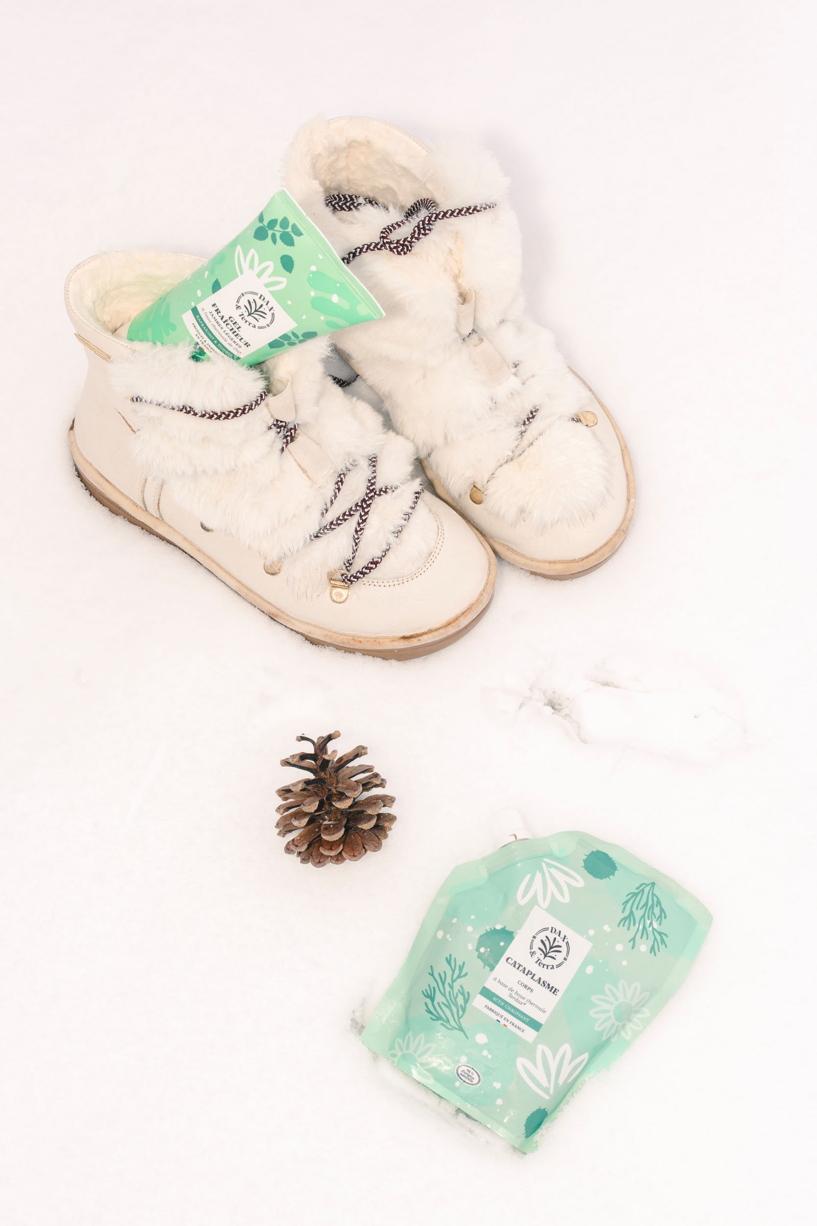 White winter boots with faux fur lining and red and white laces, a pinecone, and green herbal products on snow.