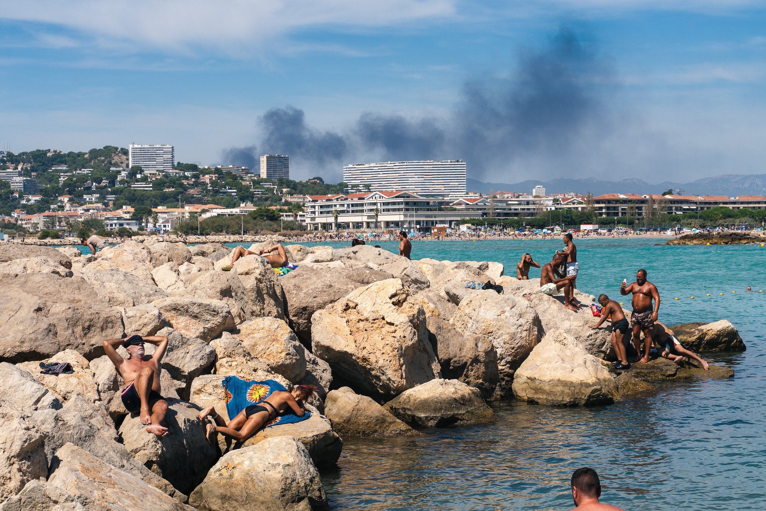 People relaxing on rocks and swimming in the ocean at a beach, with city buildings and smoke in the background.