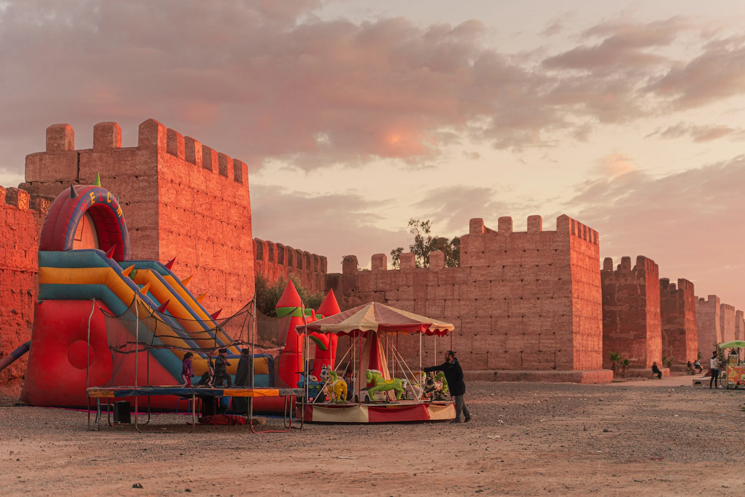 Toboggan gonflable coloré et manège avec enfants et adultes devant une ancienne forteresse en briques rouges, au coucher du soleil, à Taroudant, au Maroc