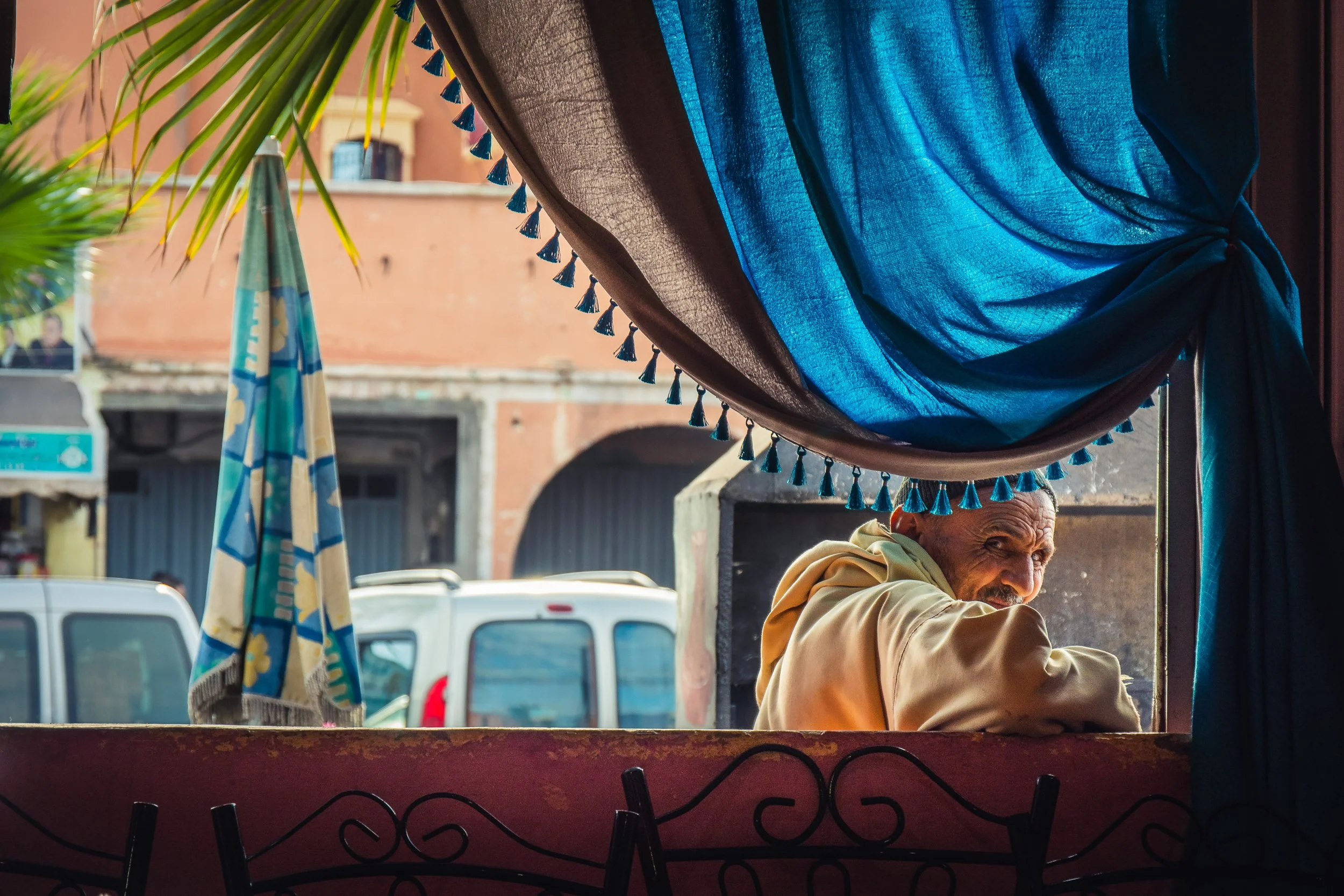 Man sitting at a street-side stall or cafe, seen through a window with colorful curtains and a patterned fence, with vehicles and buildings in the background.