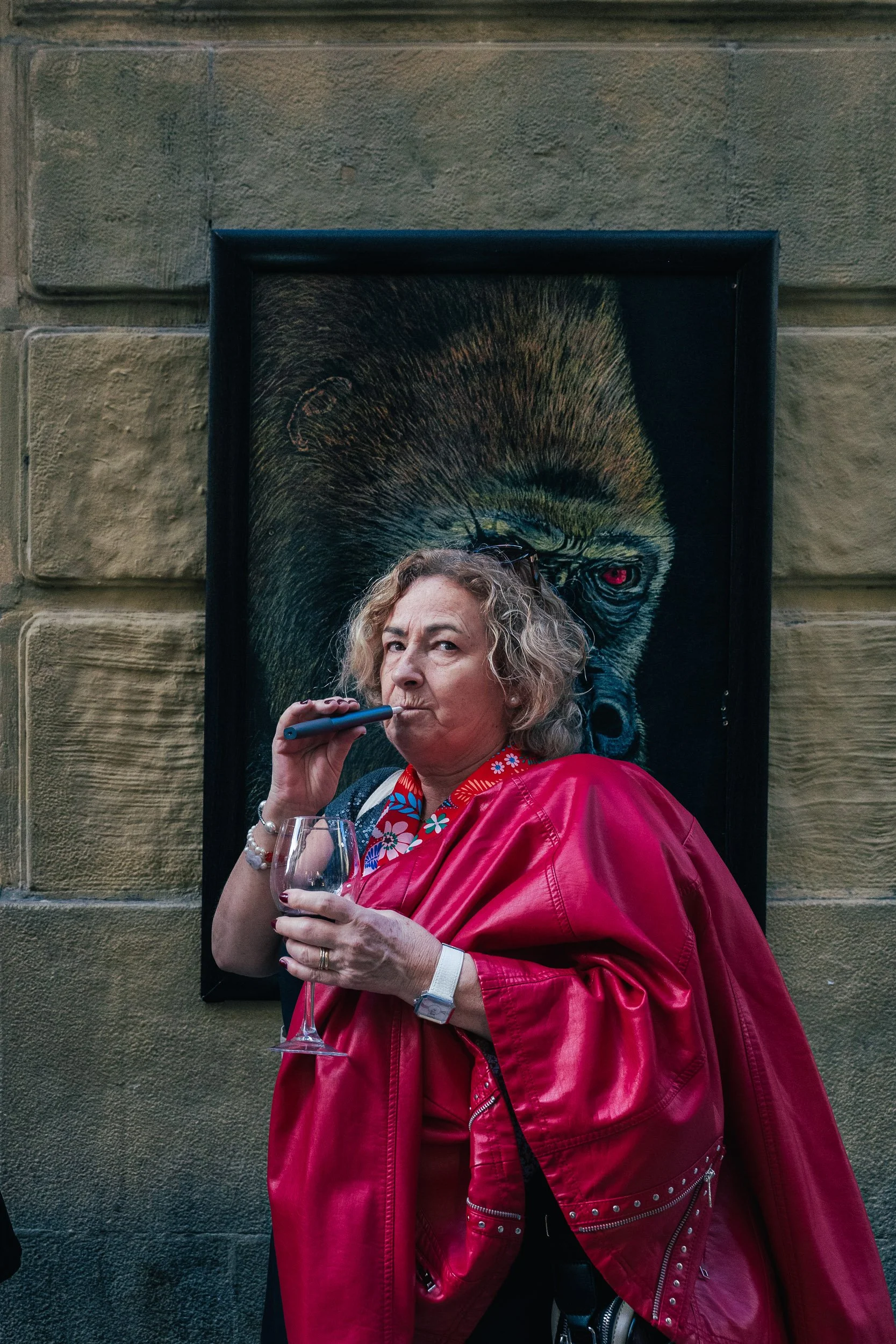 An elderly woman with curly gray hair wearing a vibrant red leather jacket holding a wine glass and a smoking vape, standing in front of a wall art depicting a gorilla with red eyes.