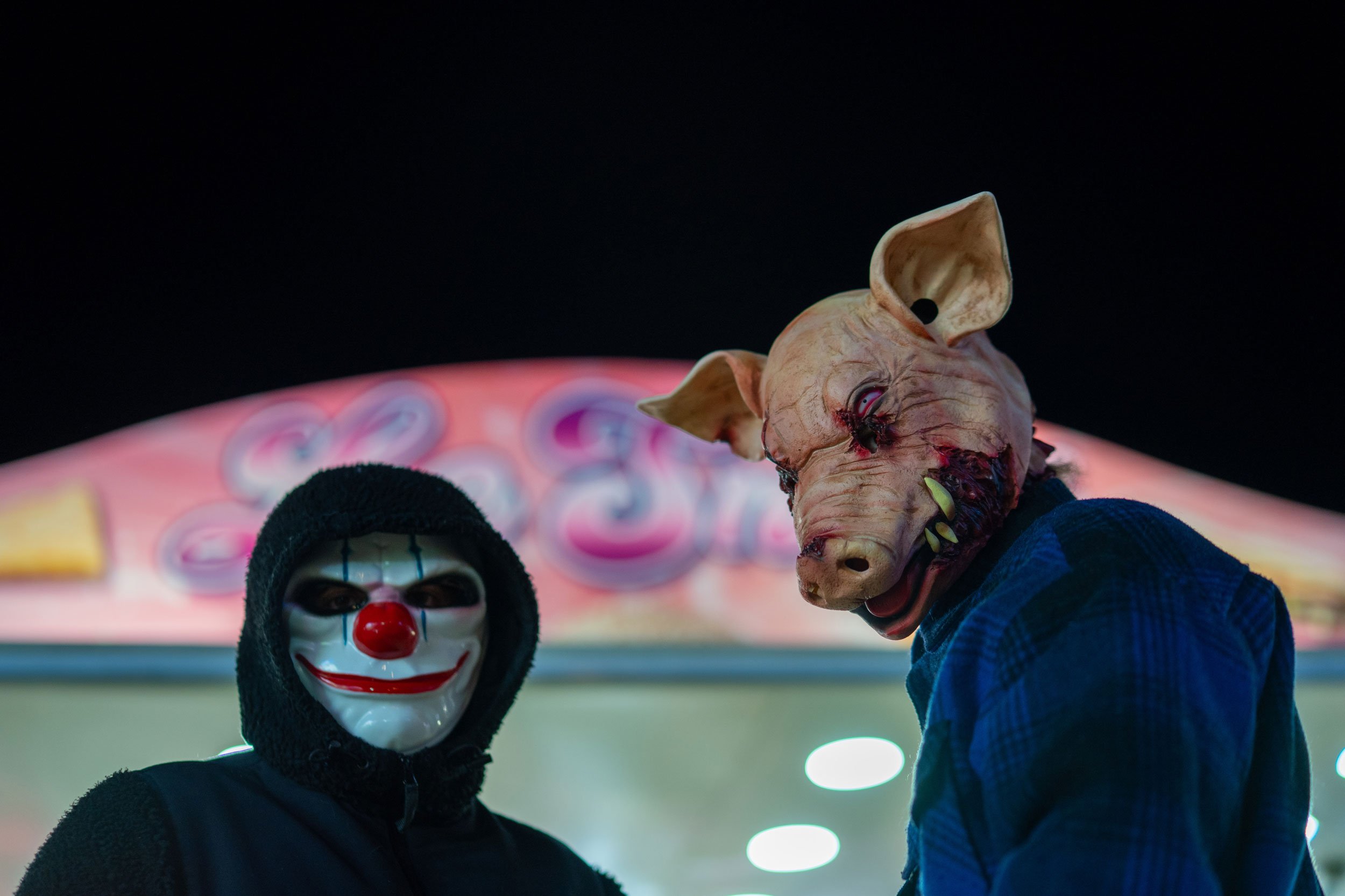 Two people wearing creepy masks: one with a clown mask with a red nose and black hood, and the other with a pig mask with missing teeth, red eyes, and ears, against a carnival or amusement park background at night.