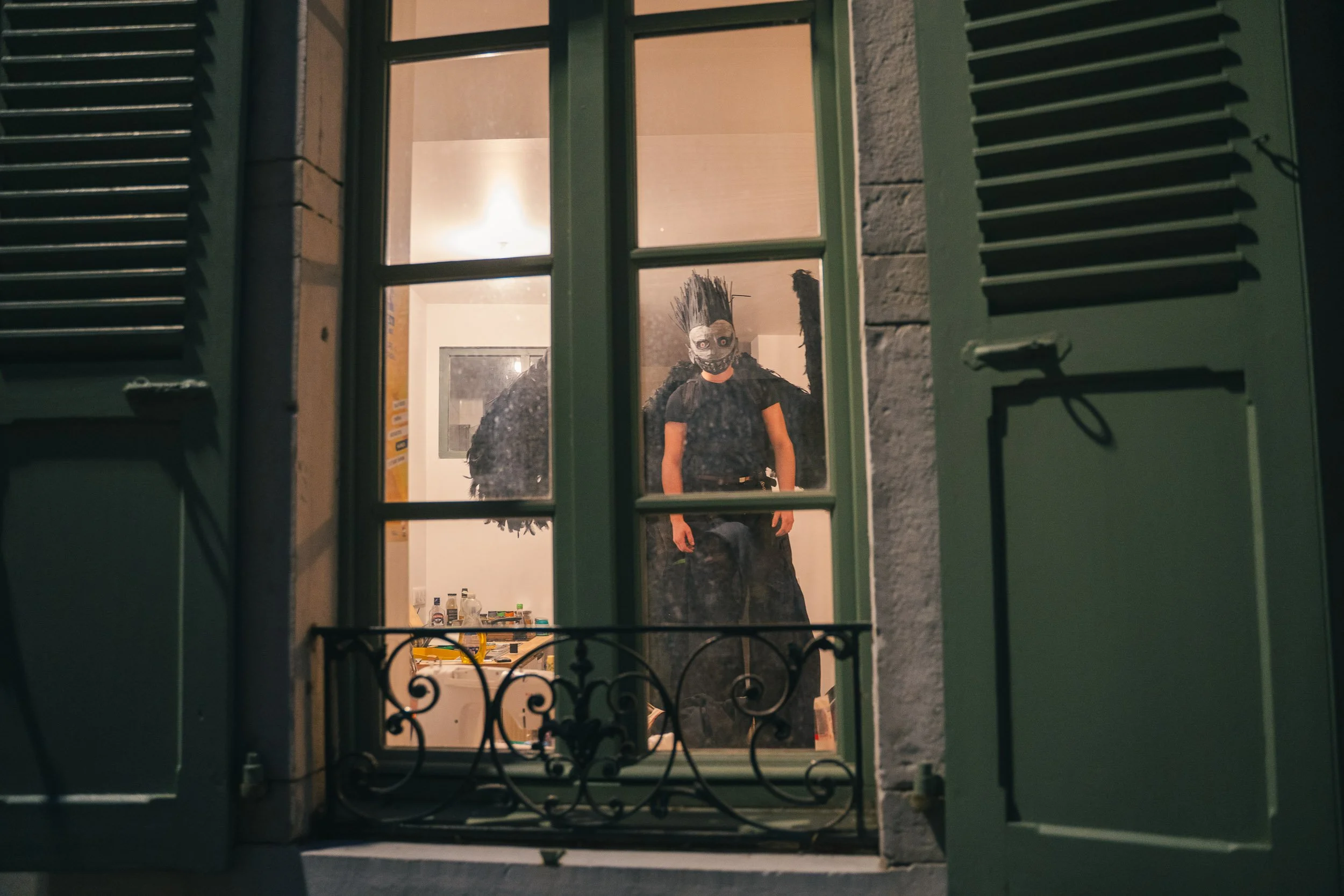 Person dressed in Halloween costume with mask and wings seen through a window from outside at night.