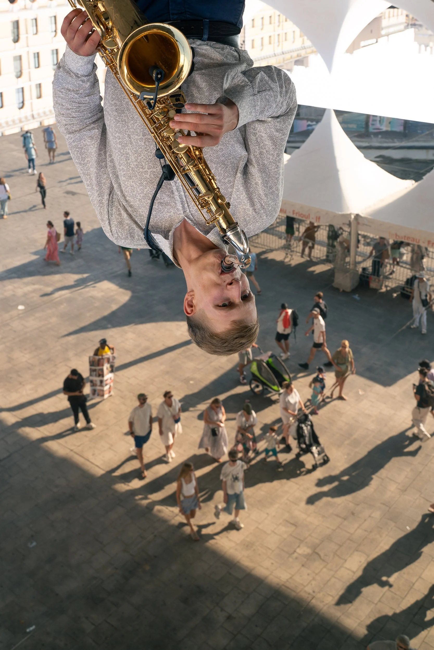 A young man playing a saxophone outdoors in a city square, with people walking below and buildings in the background.
