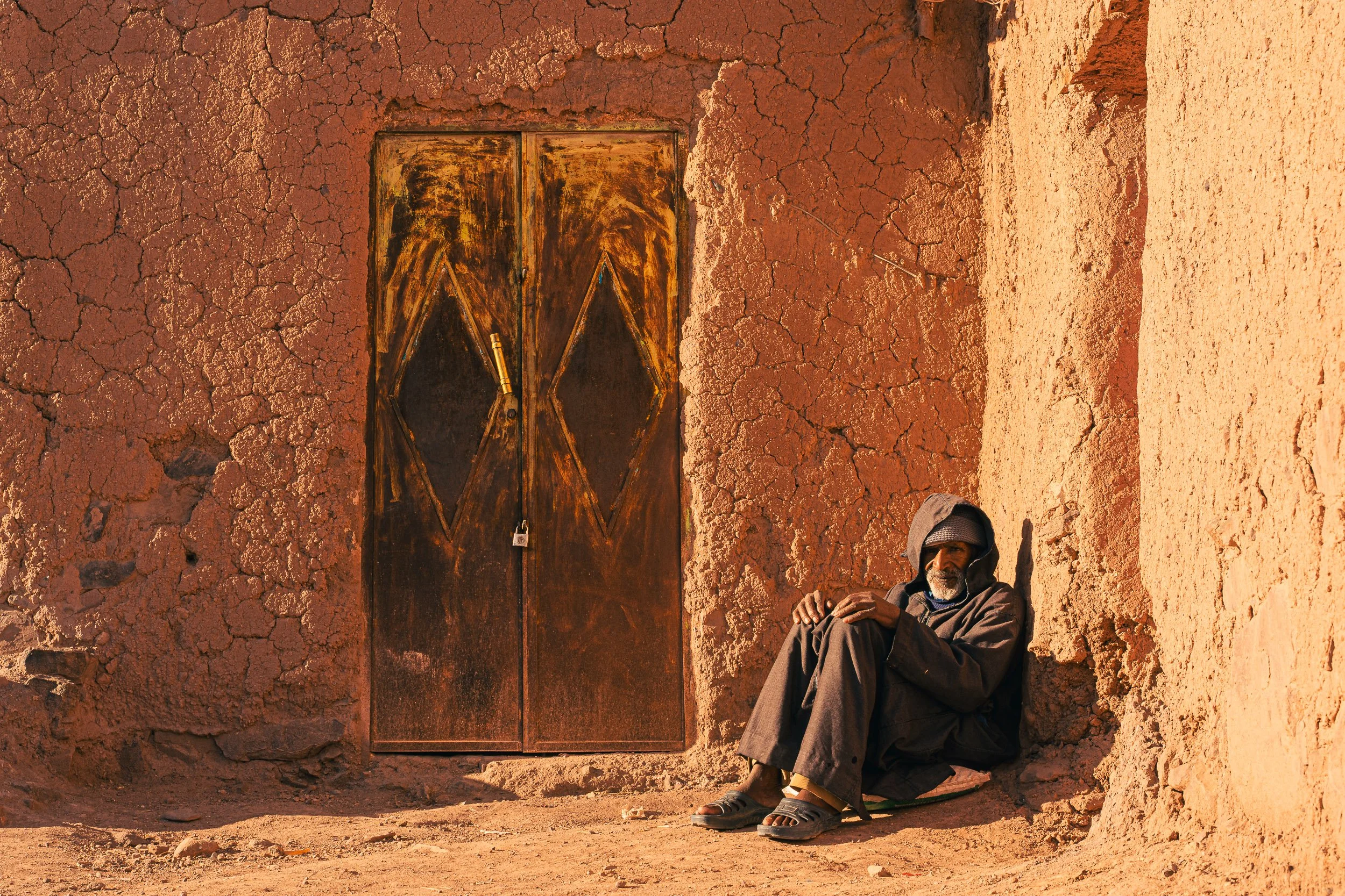 An elderly man with a gray beard sitting against a wall made of reddish mud bricks, wearing a hooded jacket and sandals, with a large metal door behind him.