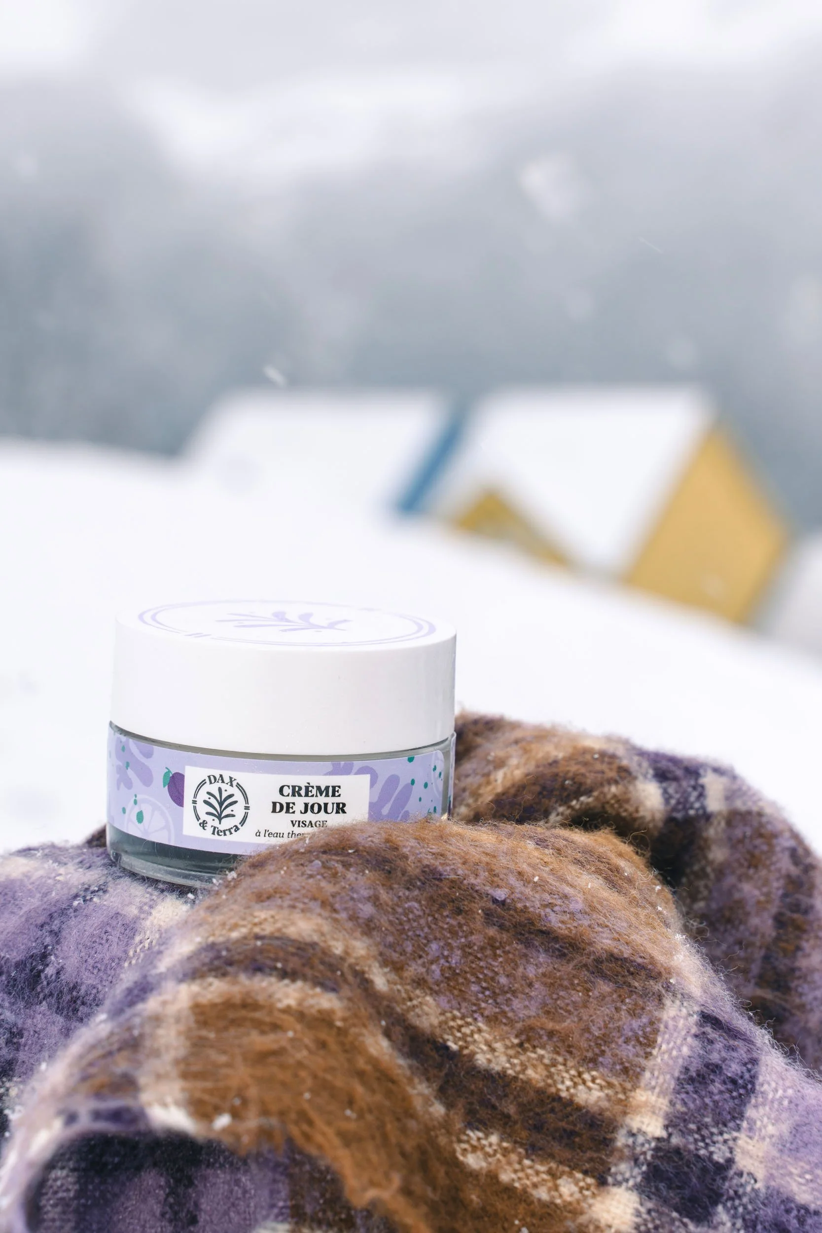 A jar of Dax & Terra face cream resting on woolen mittens in a snowy outdoors setting.
