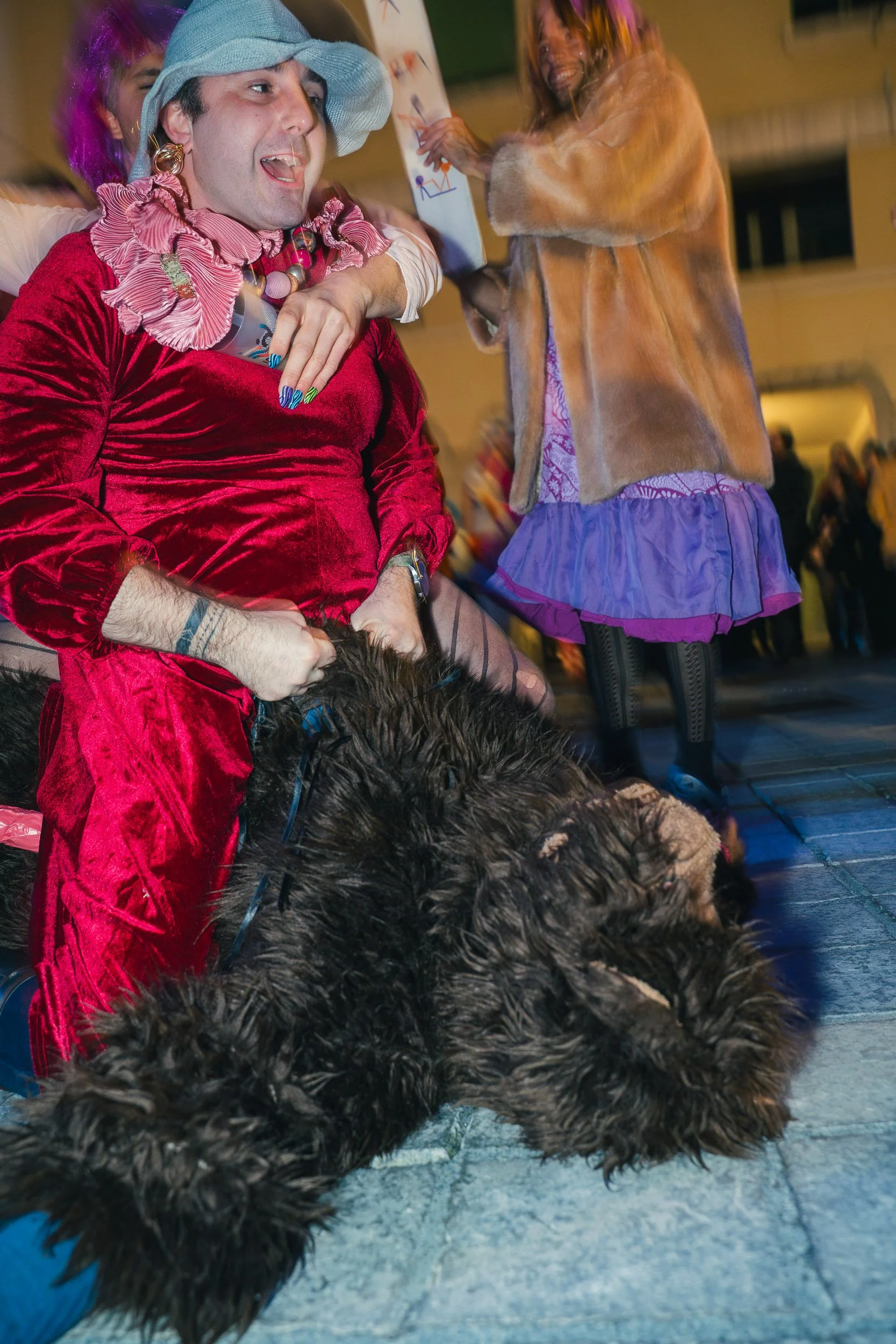Person dressed in a red velvet costume with a grey hat and colorful accessories, sitting on the ground with a large black dog lying beside them. Another person in the background is smiling and holding a paper, wearing a fur coat and a colorful skirt.