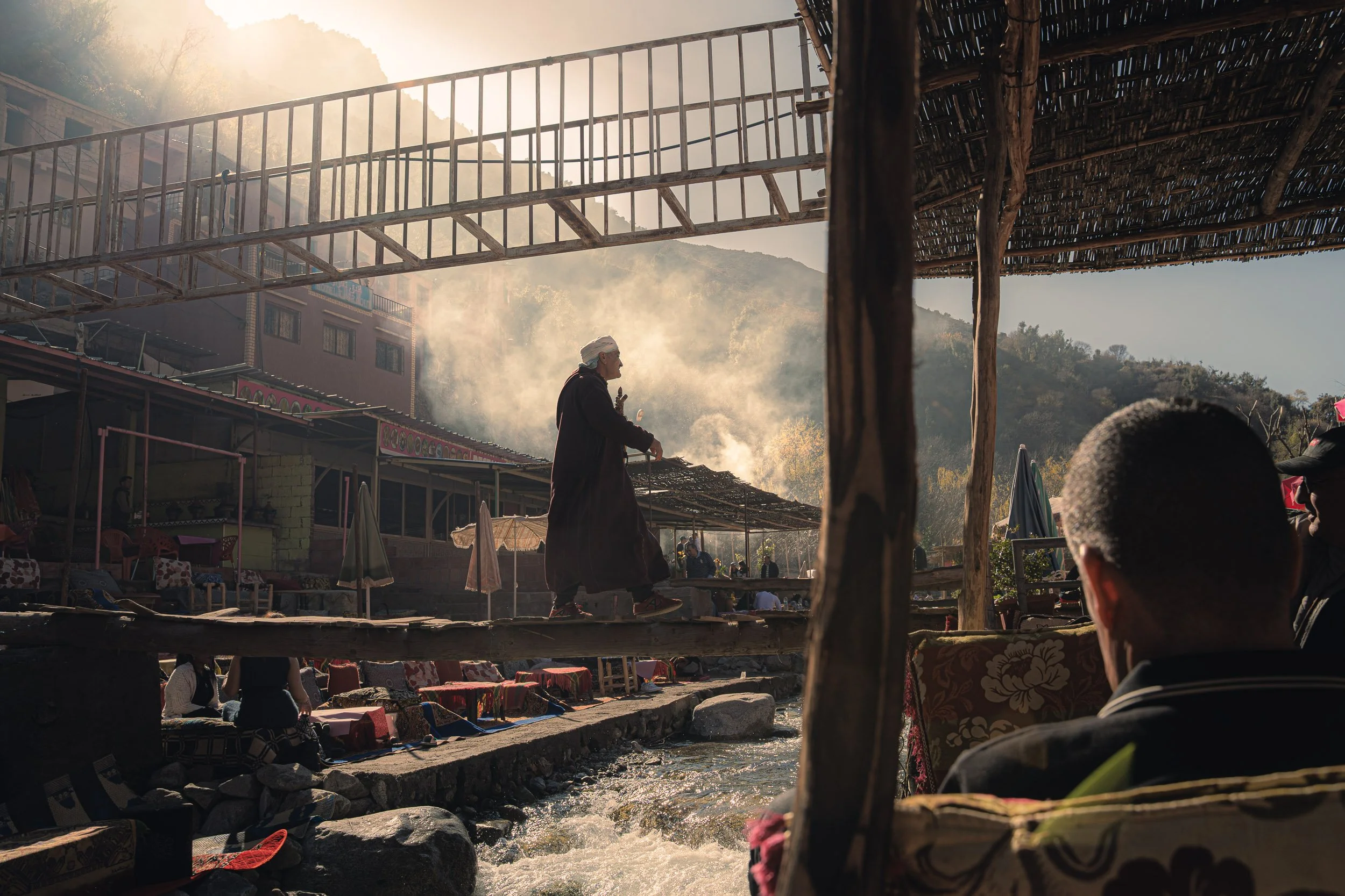A man in traditional clothing walks on a wooden bridge next to a flowing river, with several seated people nearby and a mountainous background.