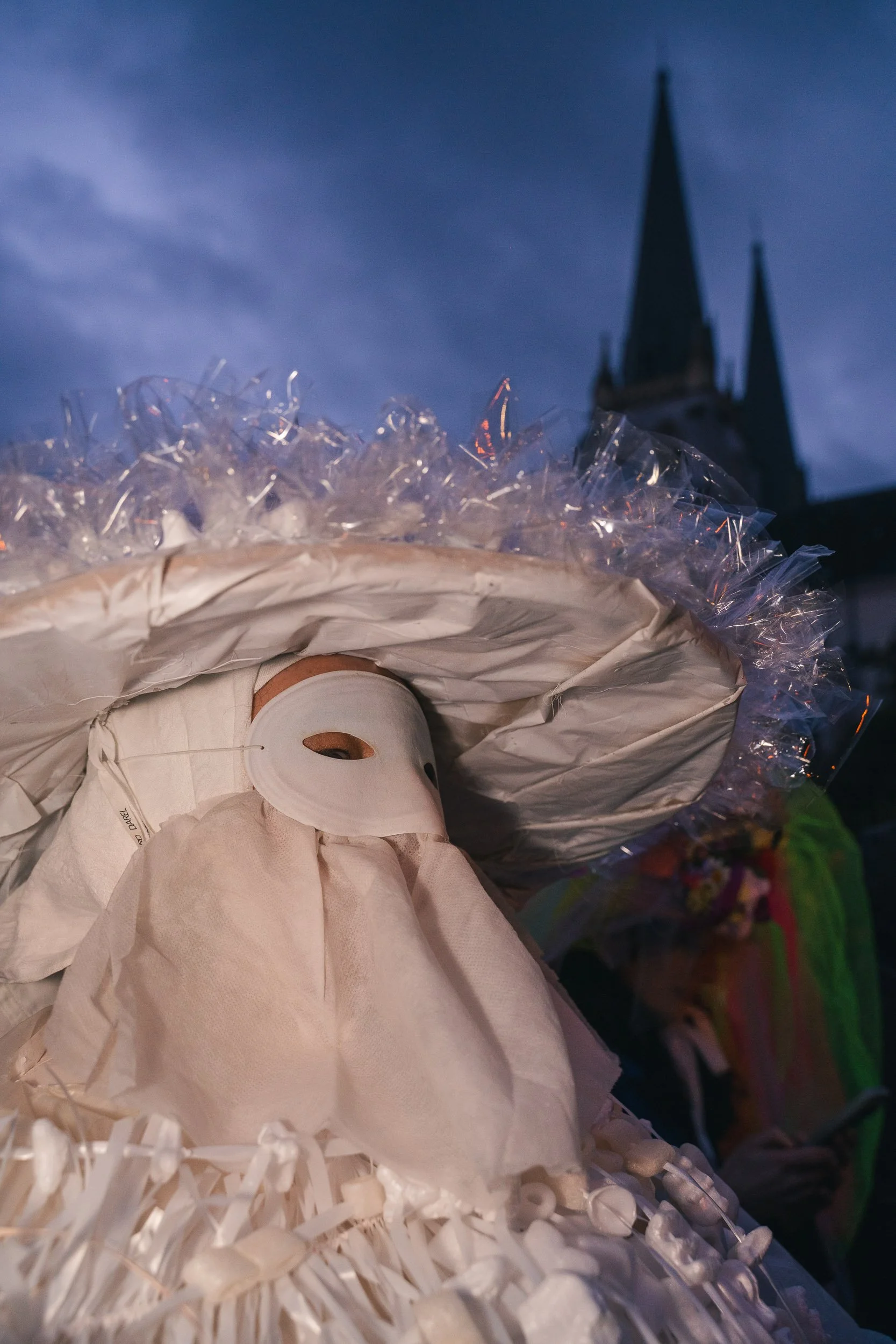 A person wearing a white mask and a large, decorative hat with iridescent trim, standing outdoors at dusk, with a church steeple in the background.