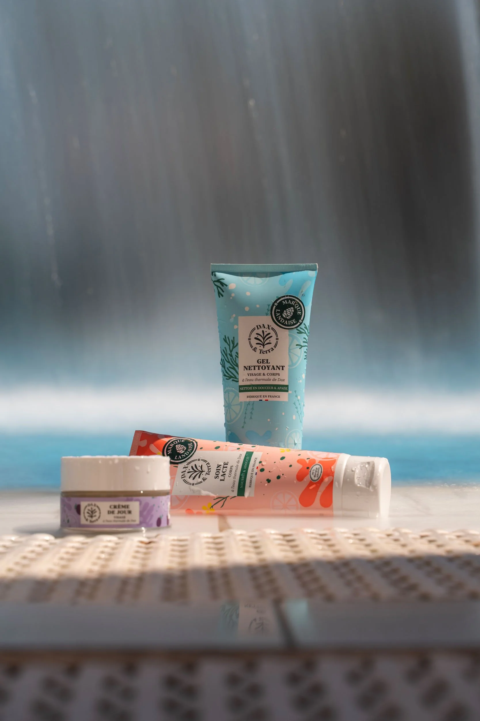 Skincare products displayed outdoors with a waterfall in the background, including a blue tube, a pink tube, and a white jar.