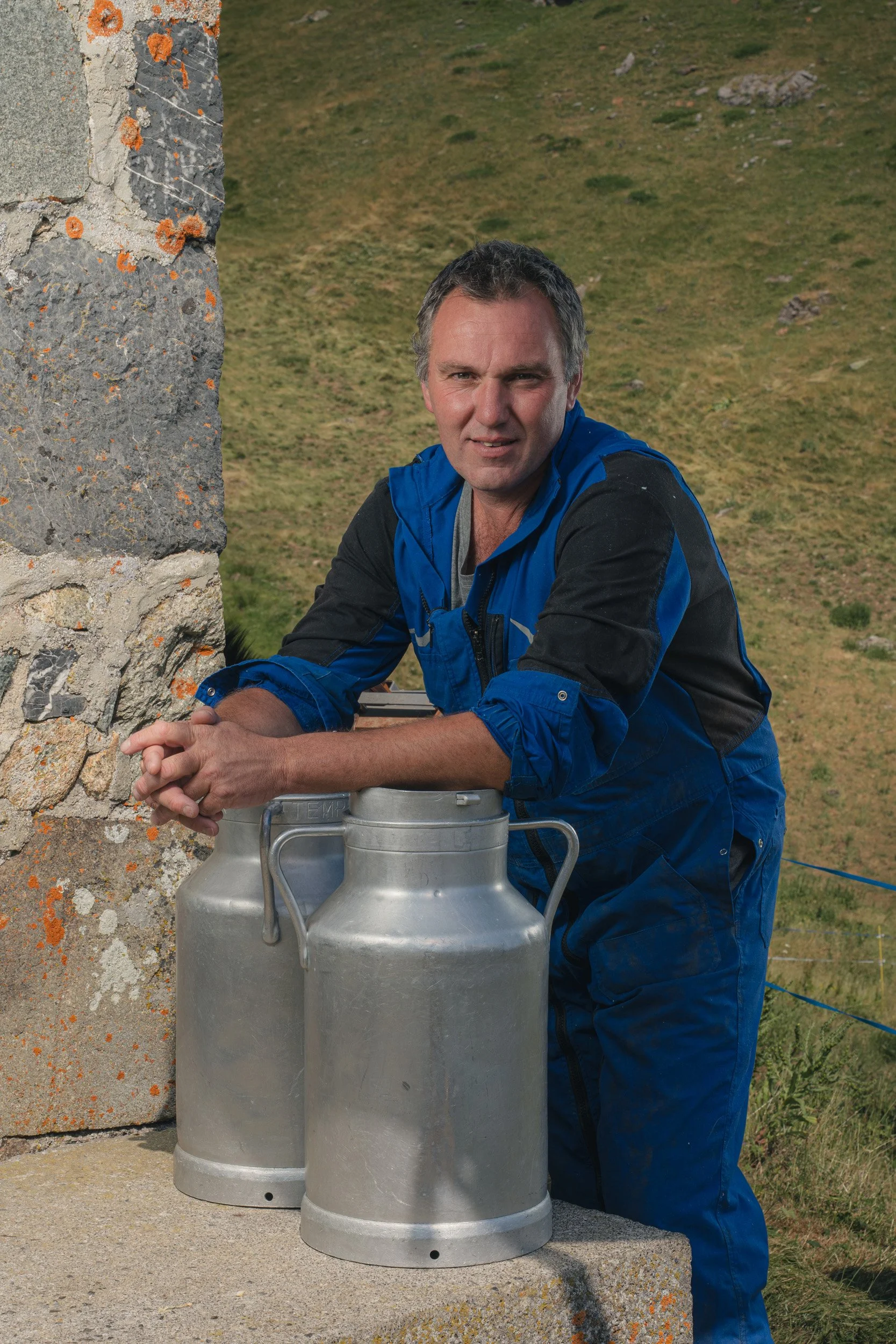 A man in a blue and black jacket leaning on a stone structure outdoors, with two large milk cans in front of him, and a grassy hillside in the background.