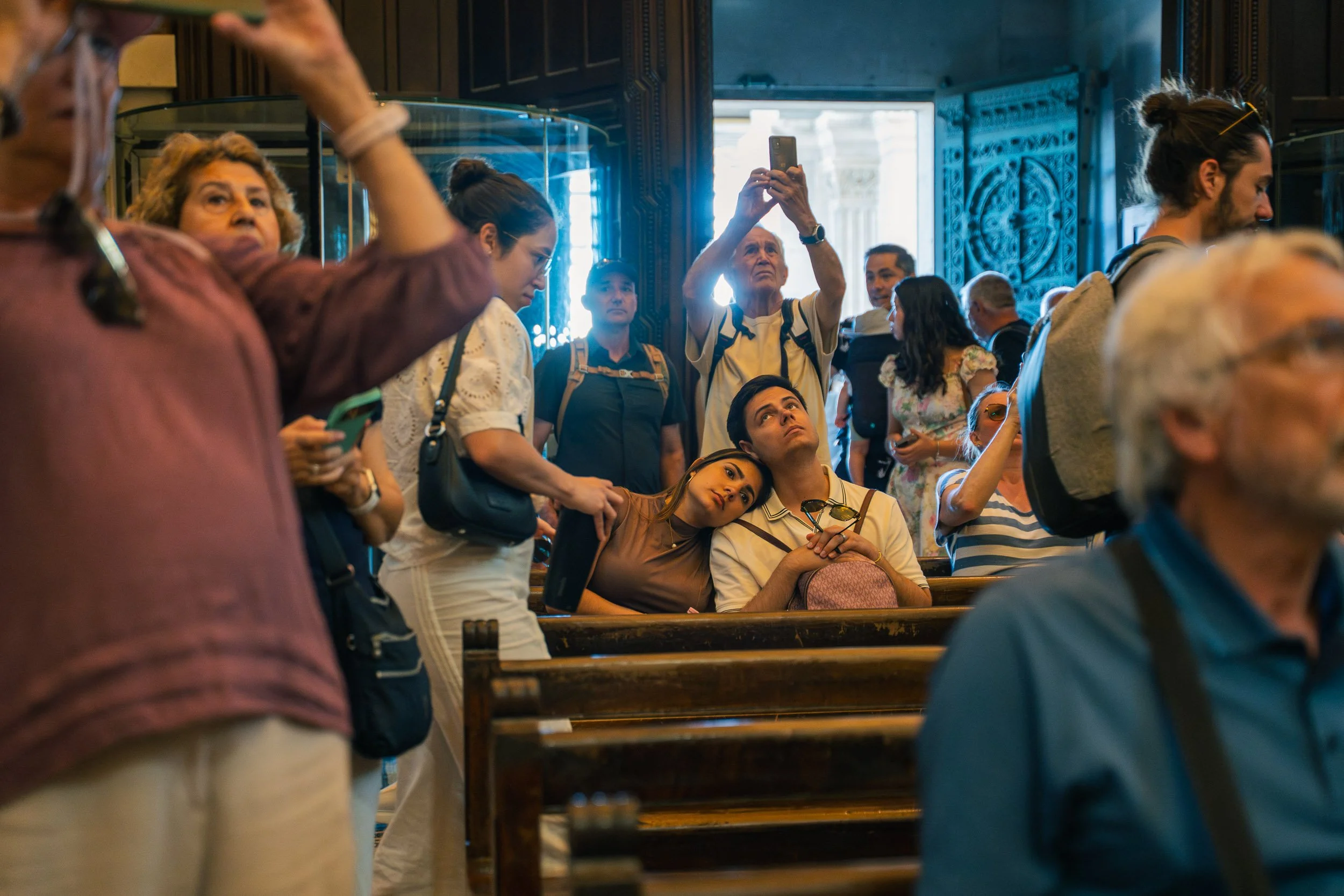 Crowded scene inside a church with people standing and sitting, some taking photos or looking around. A young woman rests her head on a young man's shoulder, both seated near the pews. An elderly man is taking a photo with his phone, and other indivi