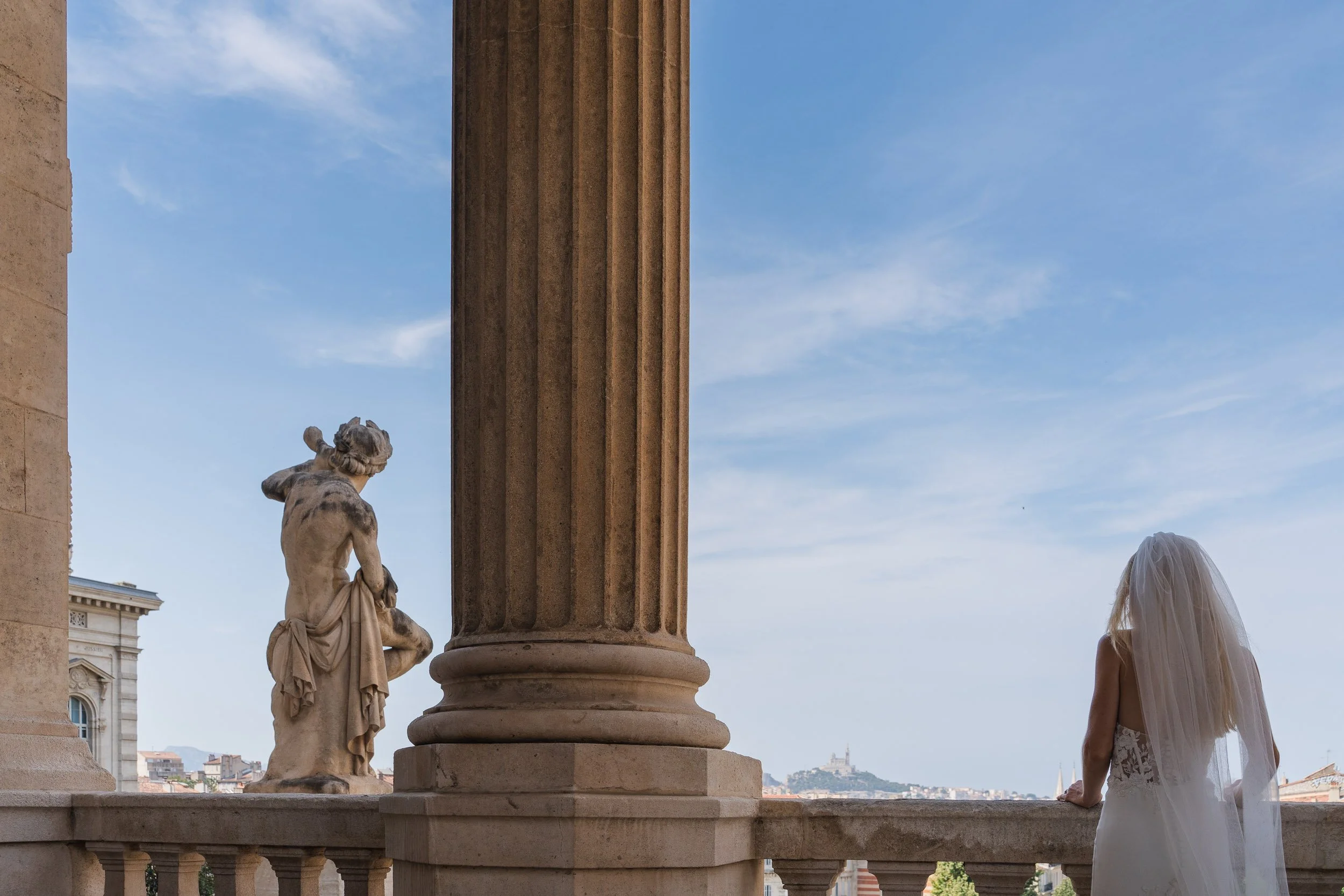 A woman in a wedding dress with a veil looking out from a balcony with classical columns and a balustrade, overlooking a cityscape with a hilltop structure in the distance.