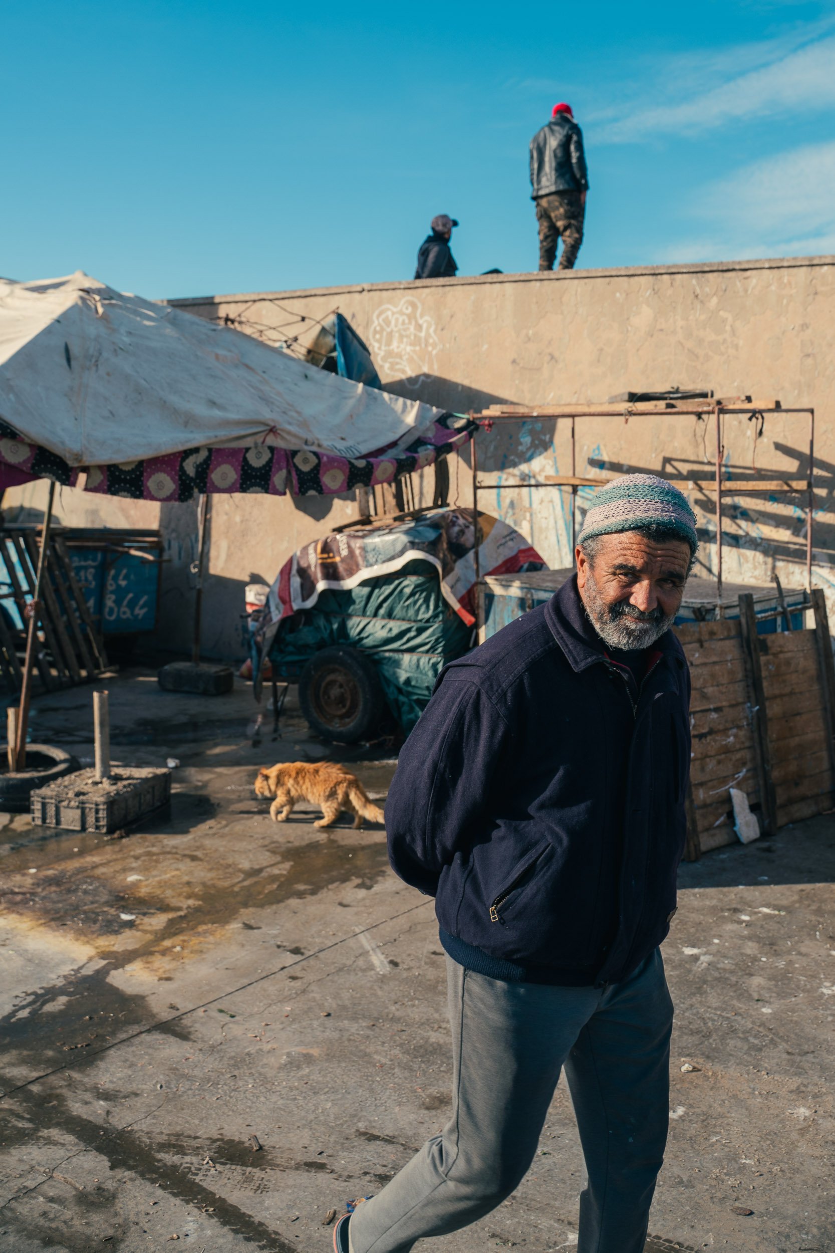 A man with a beard and gray hair, wearing a beanie and a dark jacket, standing in front of a makeshift outdoor market or shelter with a tent, a small cart, and scattered objects. Two people are walking on a wall in the background, and a small dog is 