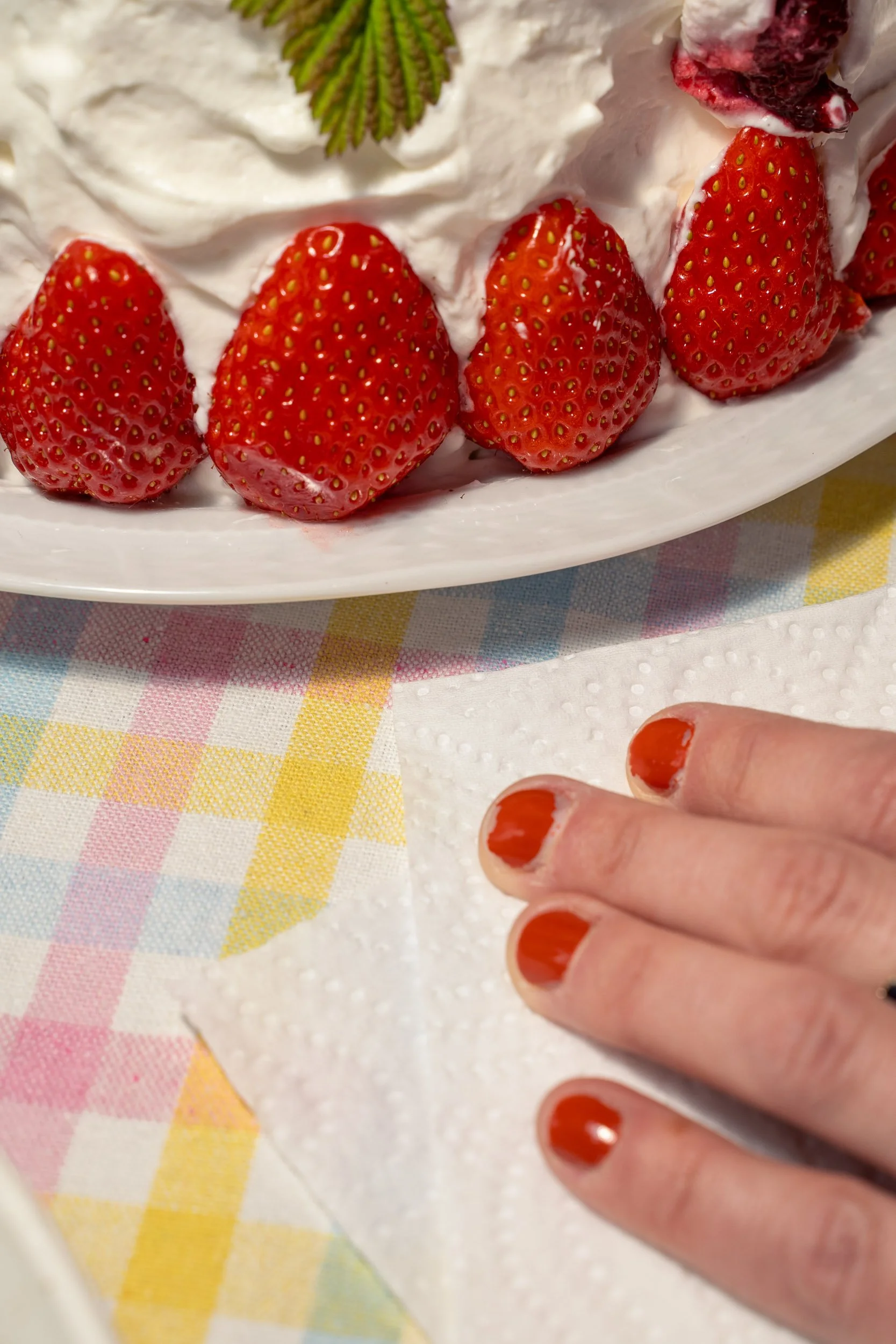 Close-up of a plate of strawberries covered with whipped cream and a mint leaf on top, next to a hand with freshly painted orange nails resting on a napkin on a pastel-colored checkered tablecloth.