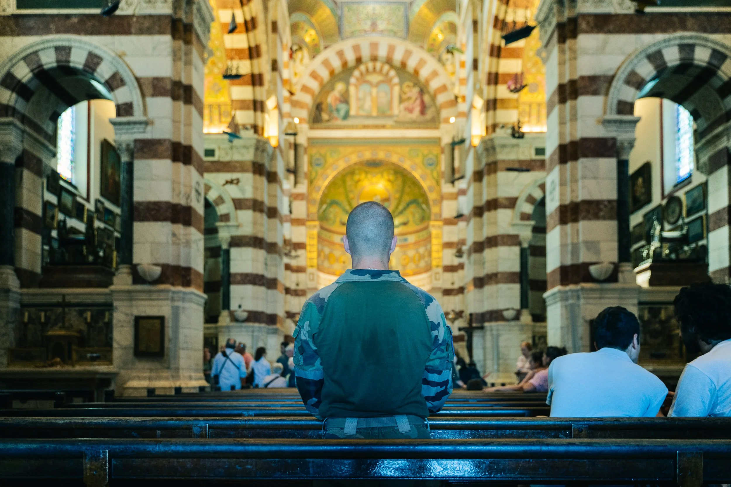 Person with a shaved head sitting in a church pew, facing an ornate altar with archways, colorful decorations, and stained glass windows.