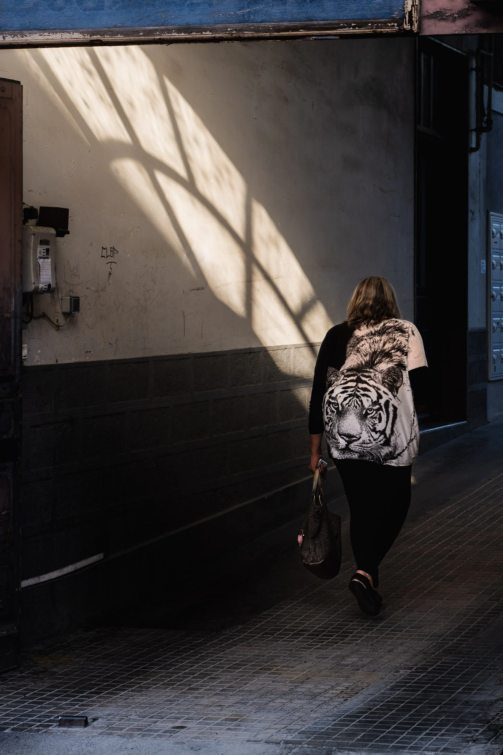 A woman walking on a city sidewalk carrying a bag, wearing a shirt with a tiger print on the back, with shadows and light patterns on the wall nearby.