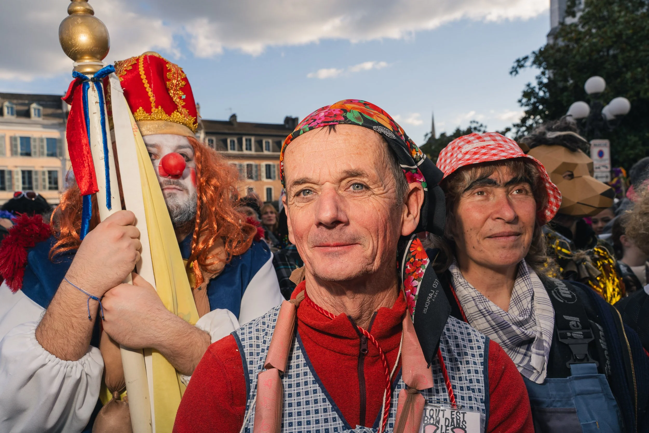 Group of people dressed in costumes, including a clown with a red wig, a man with a bandana, and a woman with a checkered headscarf, standing outdoors during daytime.