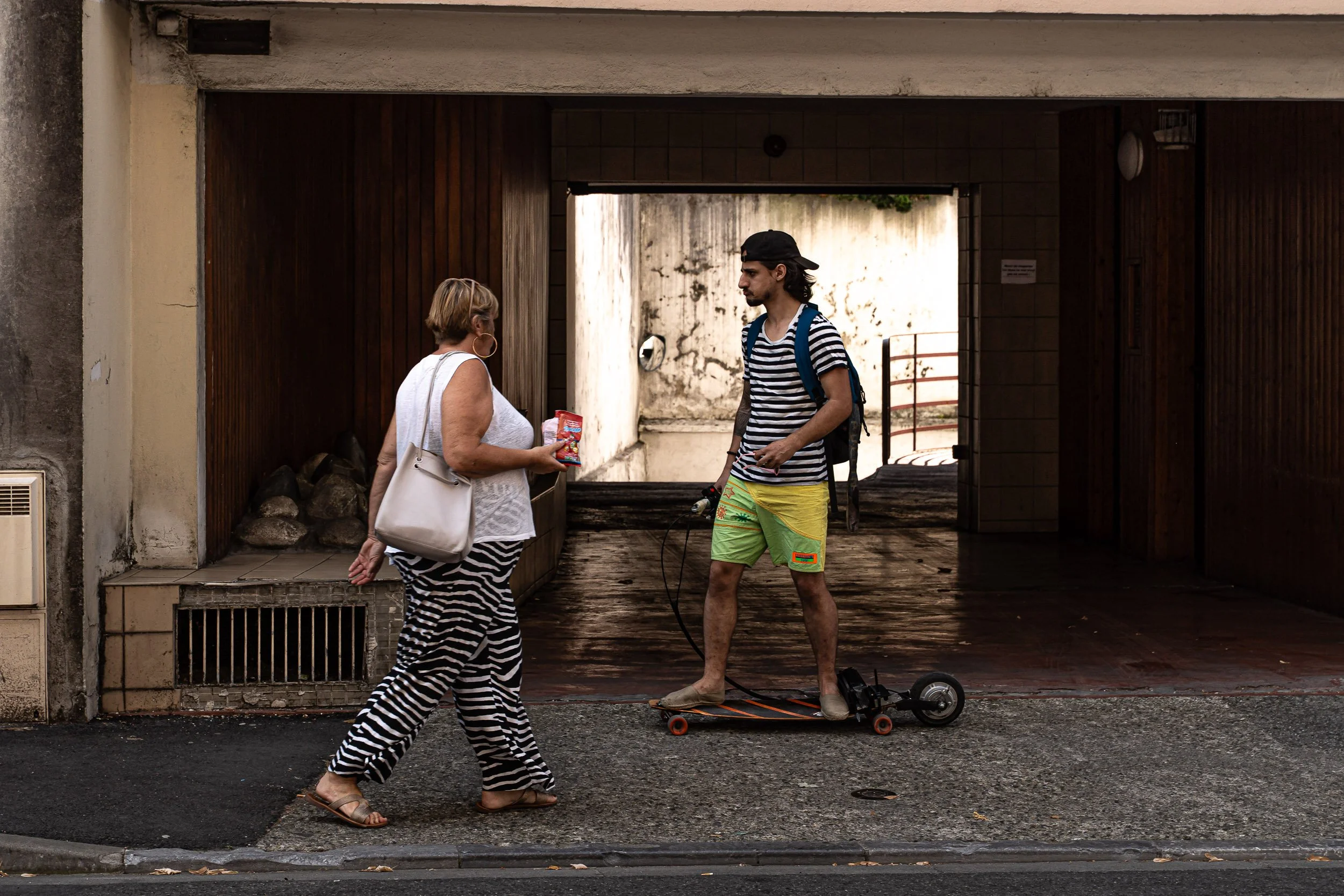 A woman walking and talking to a young man on a skateboard at an urban street scene. The woman is wearing a white sleeveless top, black and white striped pants, and sandals, carrying a gray purse. The young man is in a striped t-shirt, colorful short