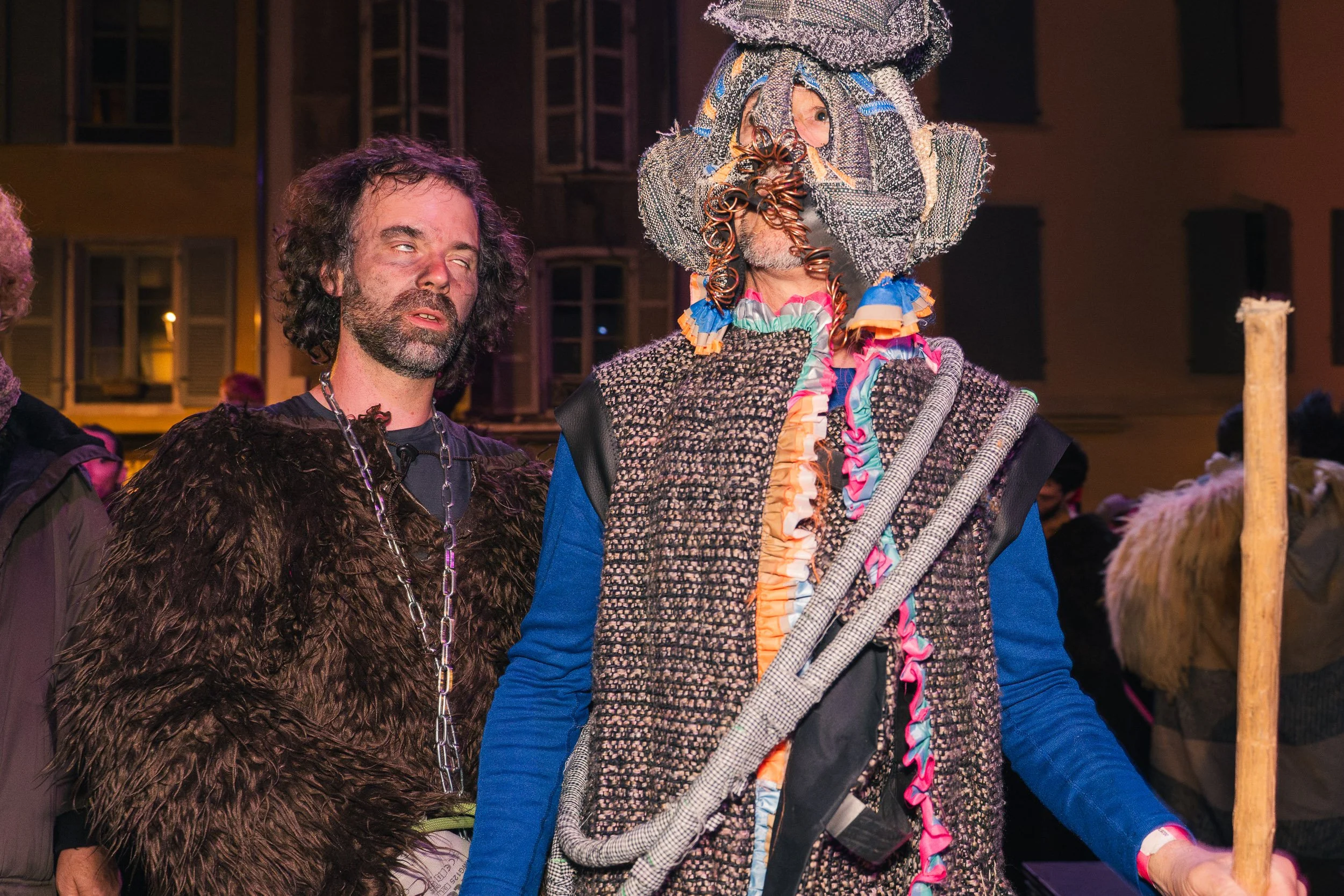 Two men in costumes, one with a beard and disheveled hair wearing a fur-like costume with a chain necklace, and the other in an elaborate, colorful, and textured outfit with a large, intricate headpiece and holding a wooden staff, at an outdoor event