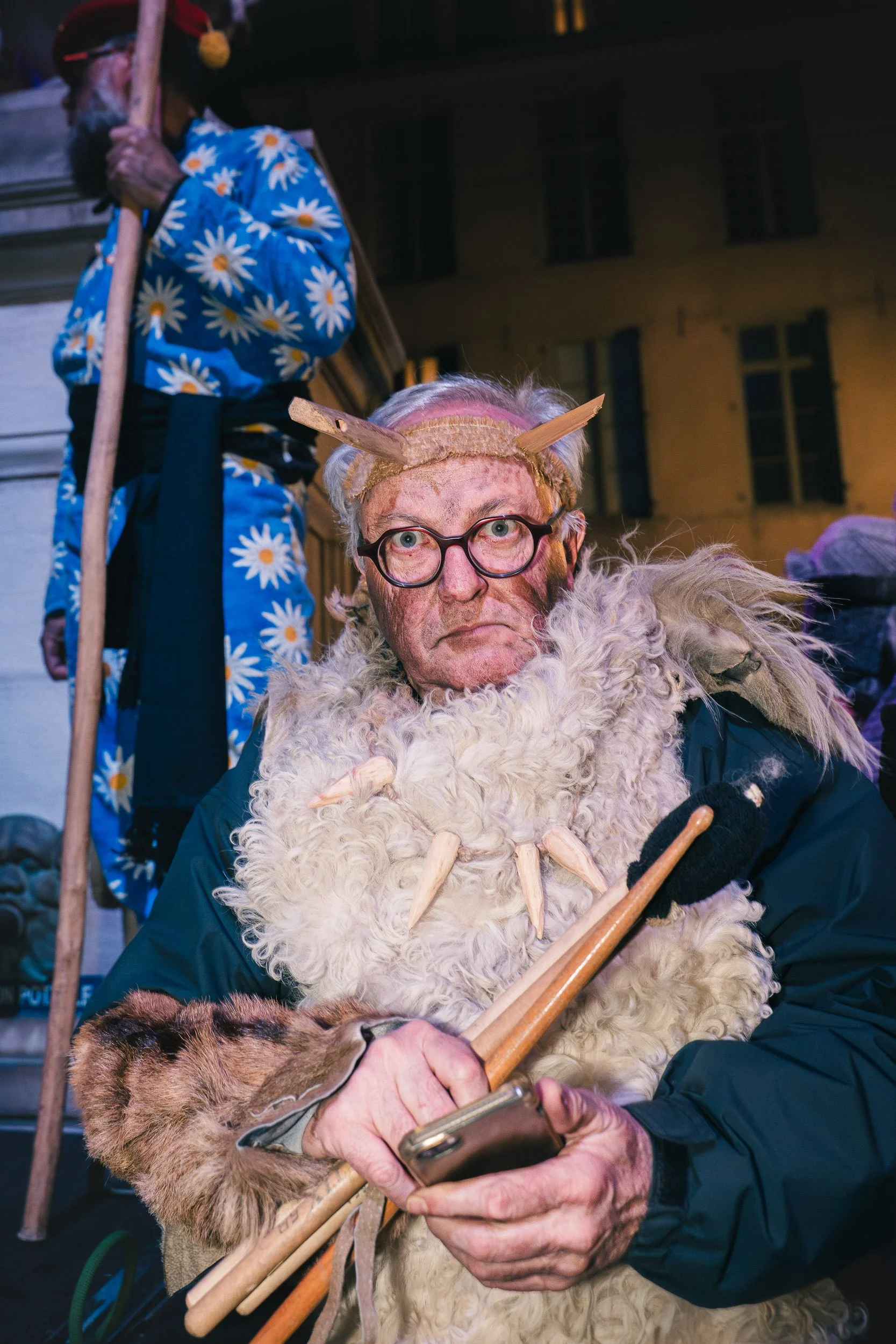 A person dressed as a mythical creature with a furry costume, horns on a headband, and holding wooden sticks, looking at a smartphone. Another person in the background wearing a blue daisy-patterned outfit and holding a staff.