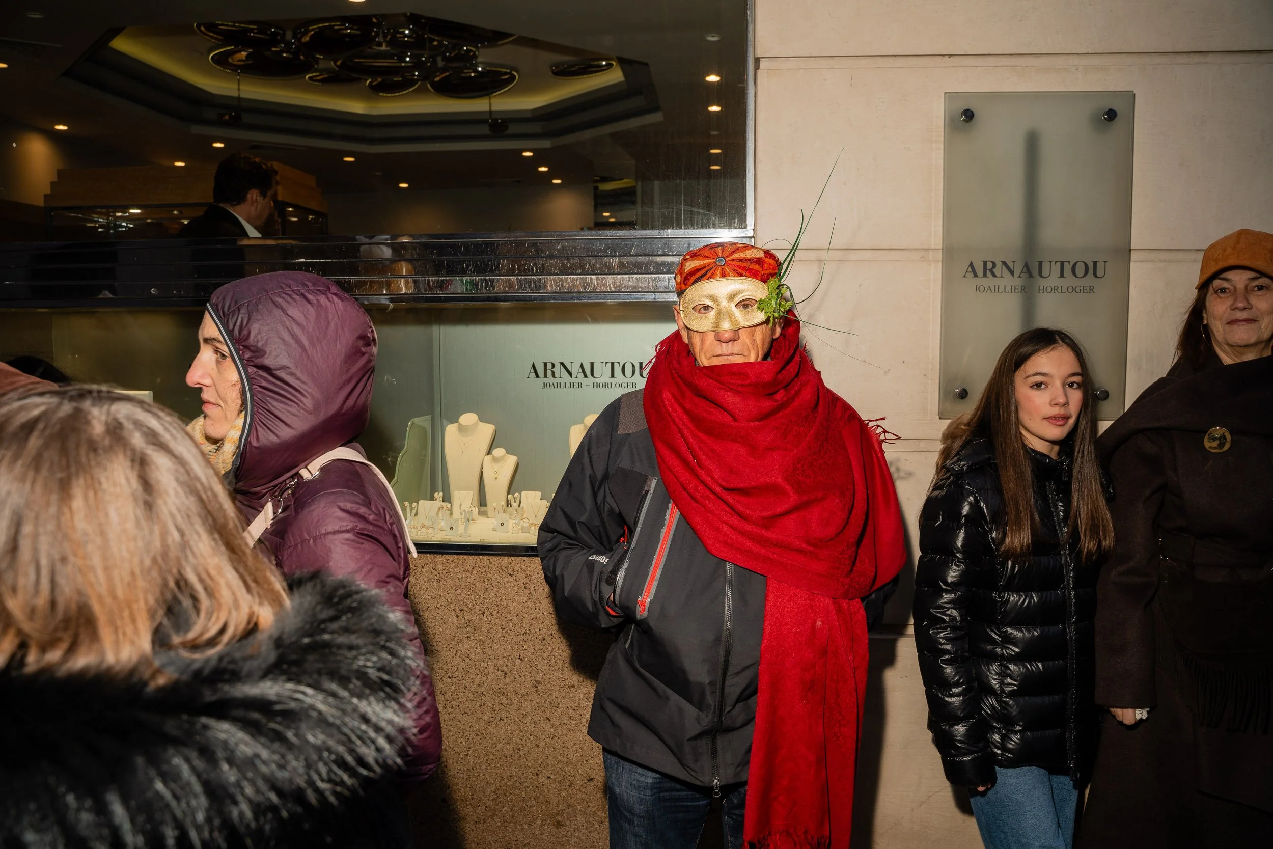 A group of people stand outside a jewelry store named Arnautou, with a display of jewelry visible through the glass window. The woman in the center wears a colorful headscarf, large decorative glasses, and a red scarf, and is looking directly at the 