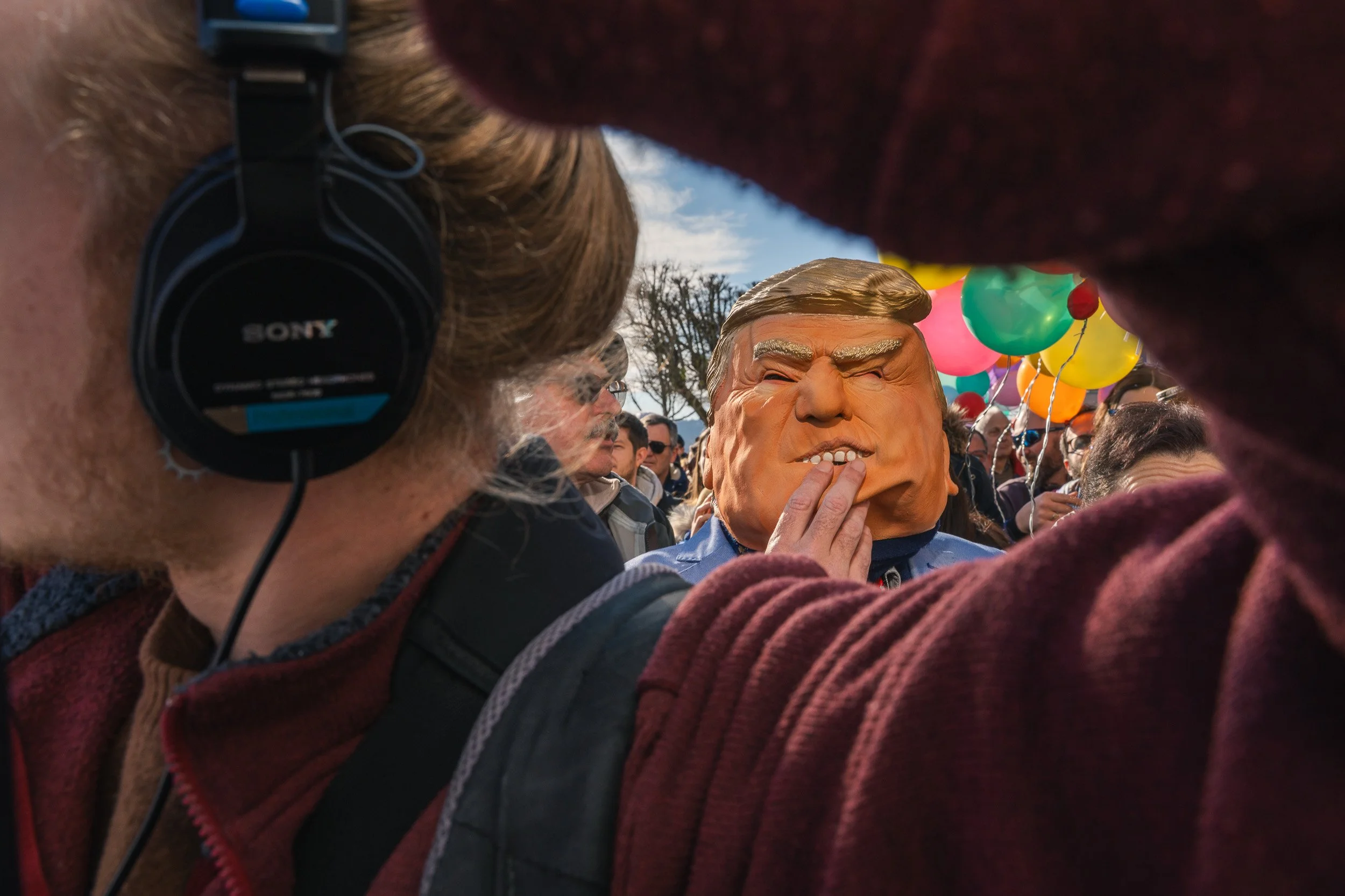 une personne qui porte un masque de Trump  et qui se tient le menton tandis qu'un perchman prend le son au premier plan, pendant le Carnaval Biarnès
