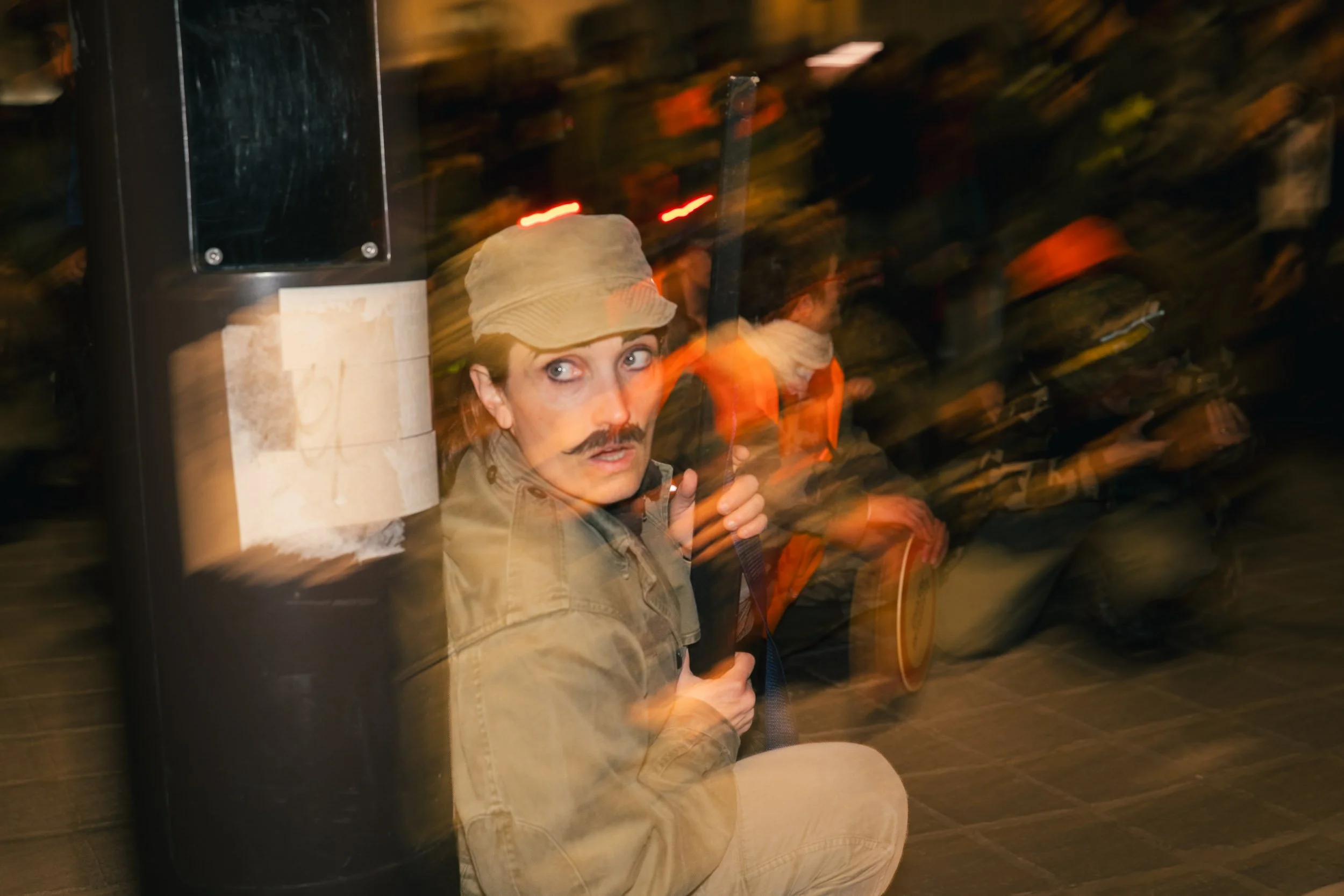 A person with a mustache and wearing a tan hat and jacket sitting on the ground at night, holding a stick or pole, with blurred figures in the background.