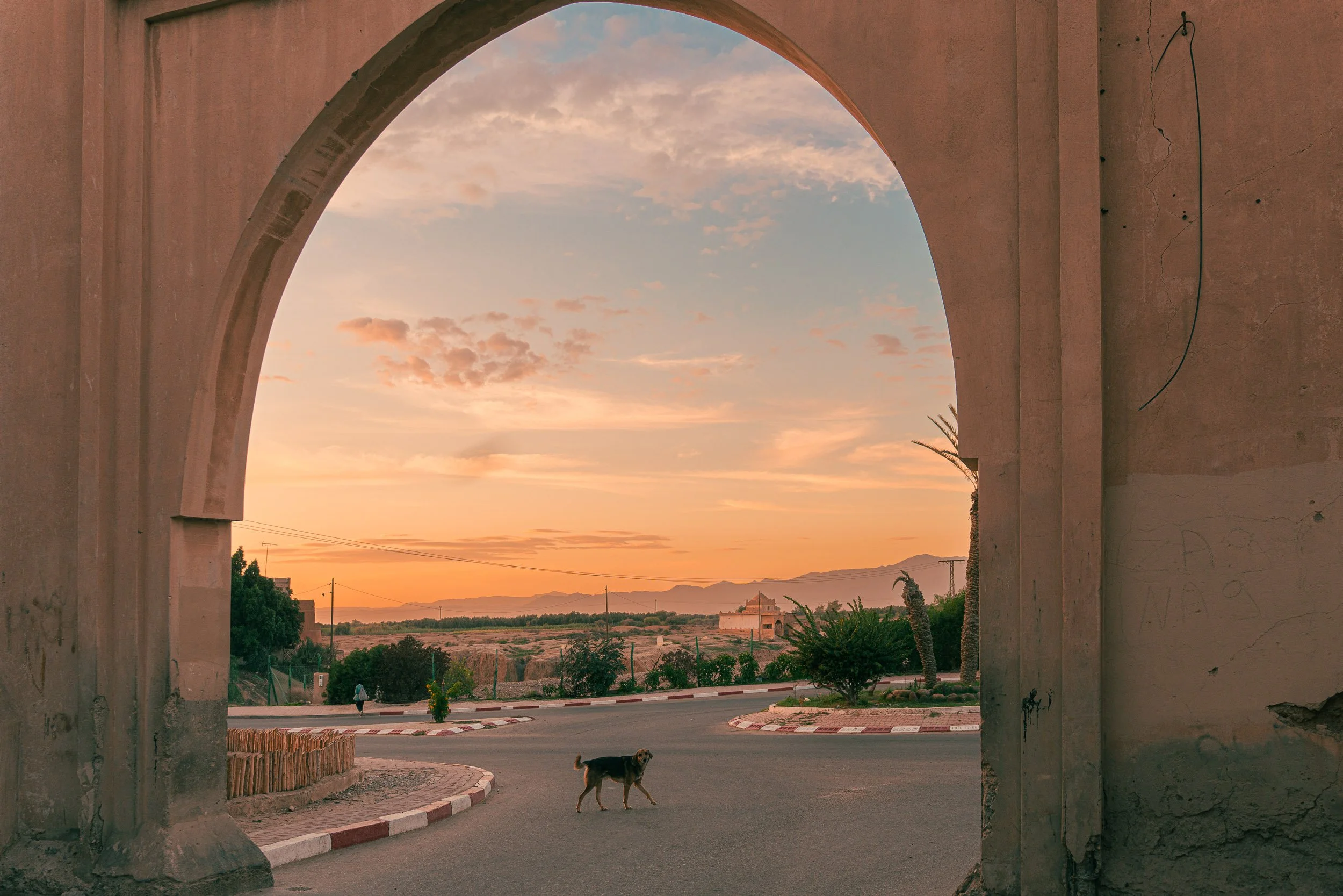 Une vue au coucher du soleil à travers une ouverture en arc, montrant une rue avec un chien et une personne, avec des montagnes au loin et un ciel partiellement nuageux au Maroc.