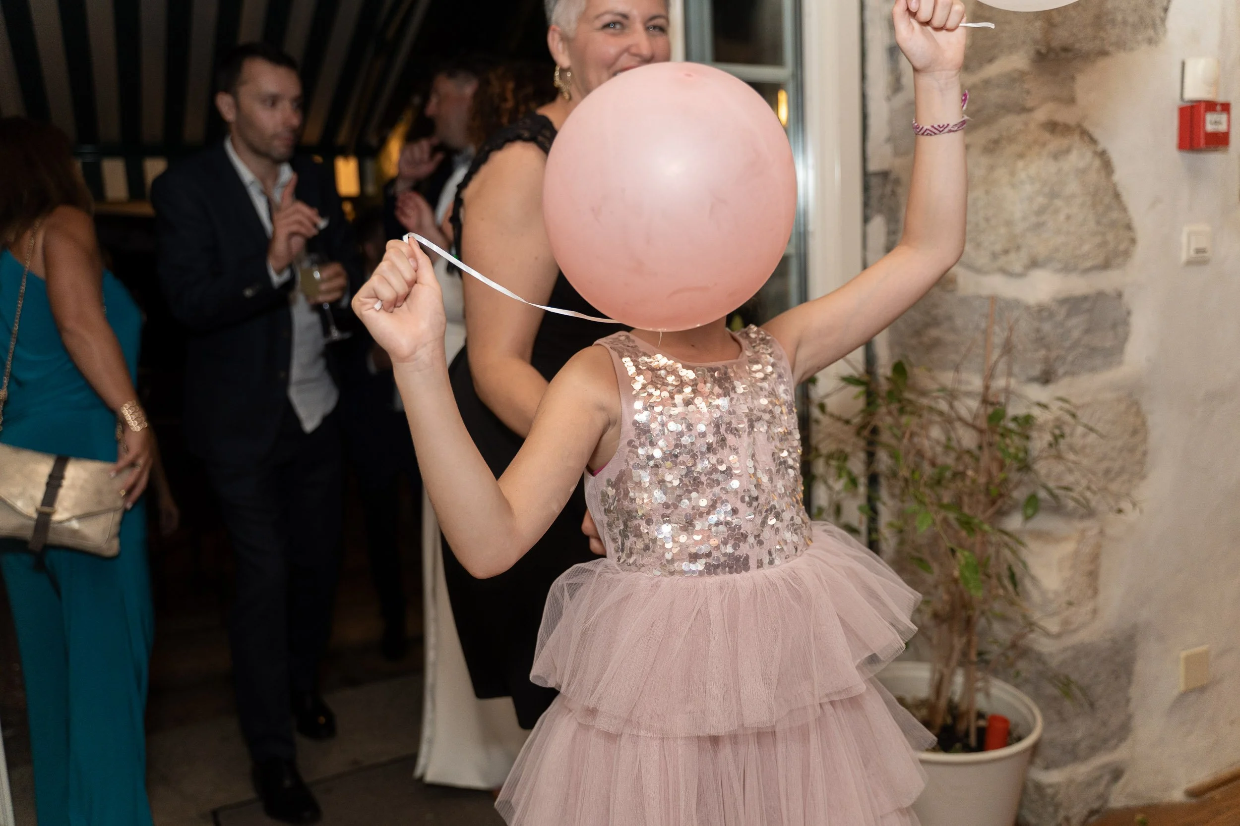 A girl in a sparkly pink dress holding a pink balloon in front of her face, at a party with adults in the background.