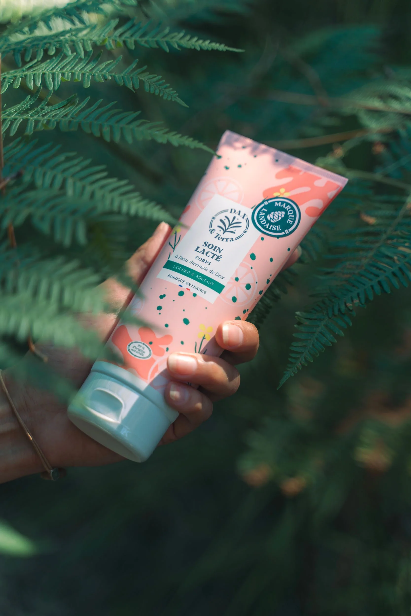 A hand holding a pink tube of body cream among green ferns.