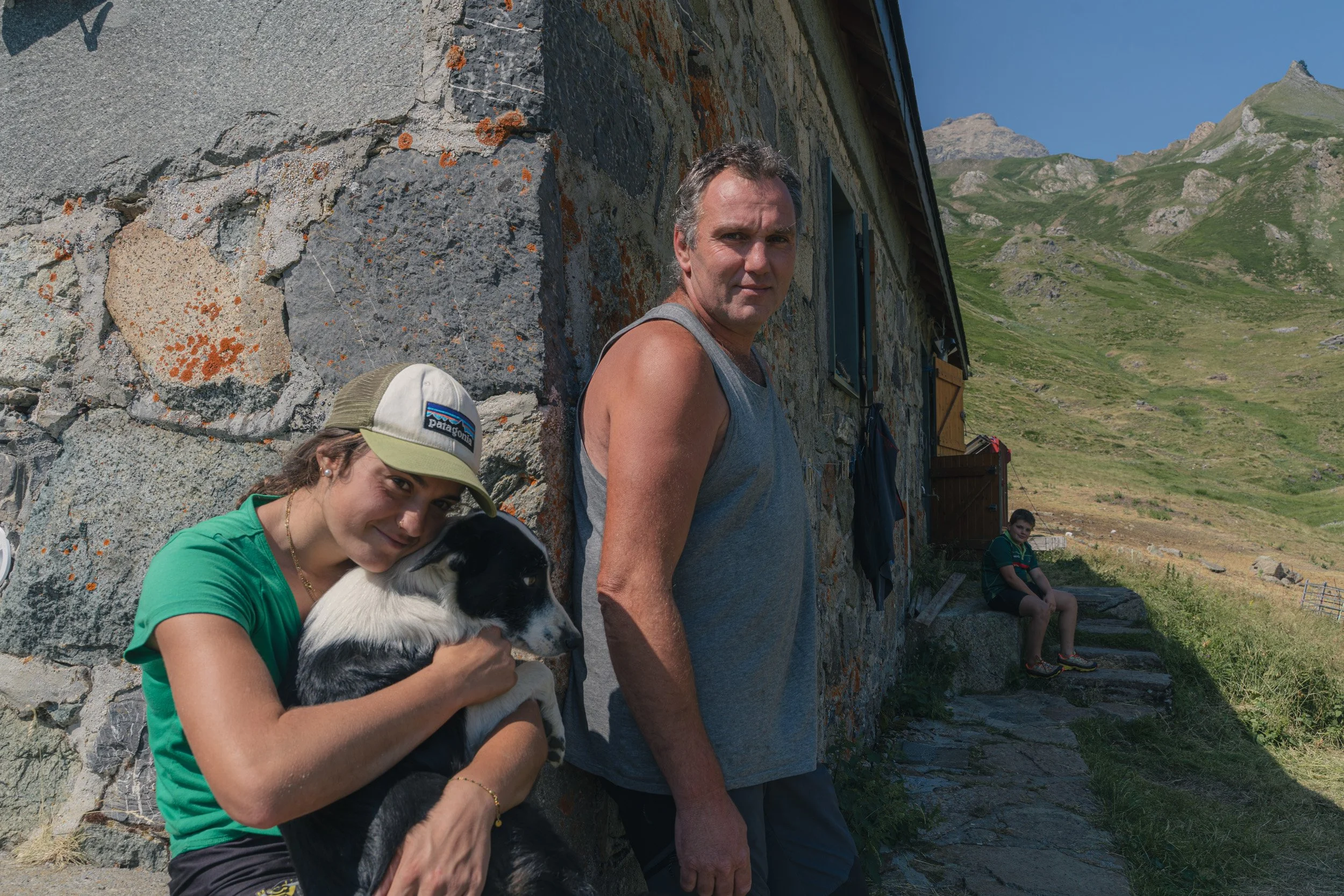 Three people and a dog outdoors beside a stone building with mountains in the background. A young woman in a green shirt and baseball cap is hugging a black and white dog. A man in a sleeveless shirt stands nearby, and a young girl sits on stone step