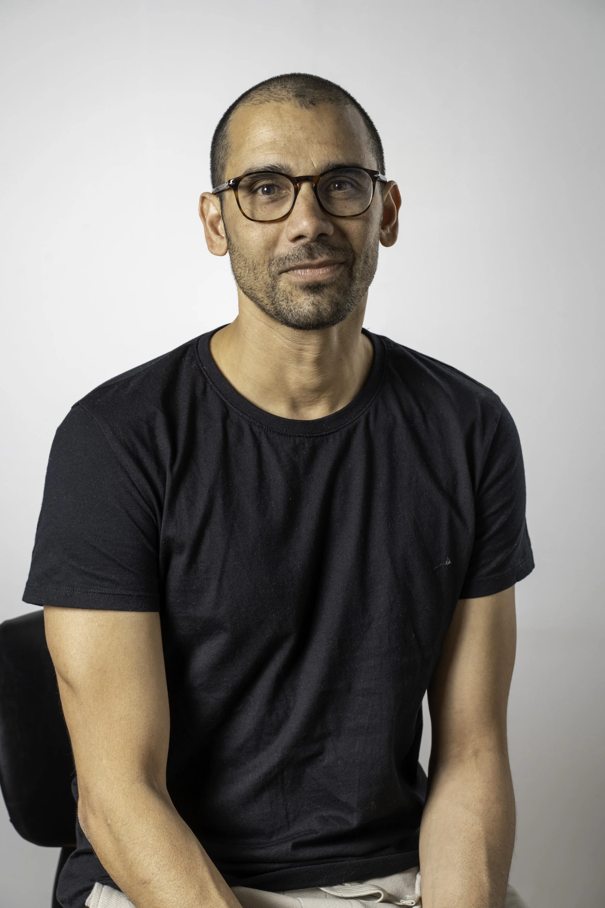 A man with short dark hair, light skin, wearing glasses and a black T-shirt sitting on a black chair against a plain white background.