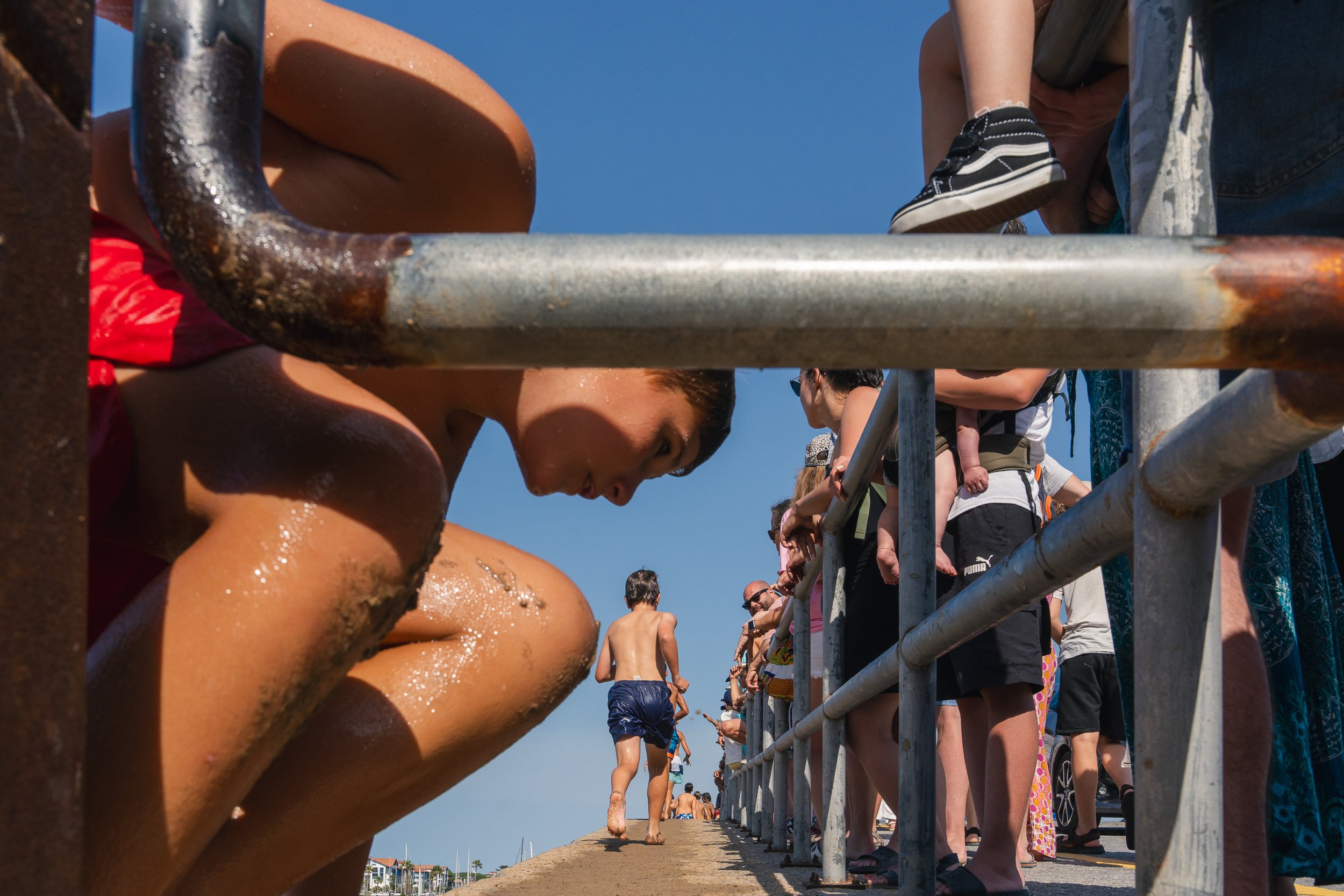 Children and adults on a pier, some wet from swimming, standing behind a metal railing during a sunny day near the beach.