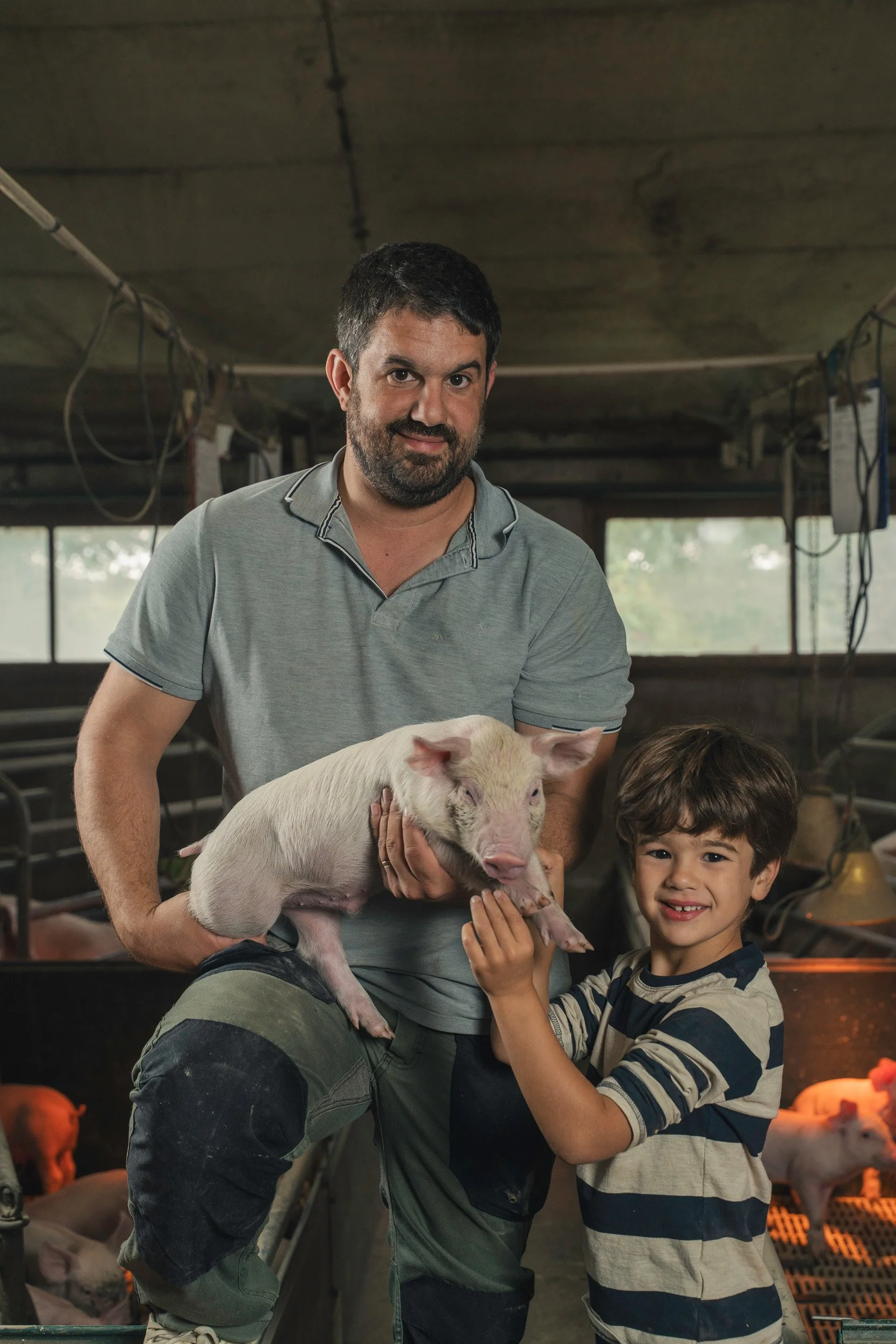 A man and two children in a pig farm holding a piglet together, with pig pens and other piglets visible in the background.