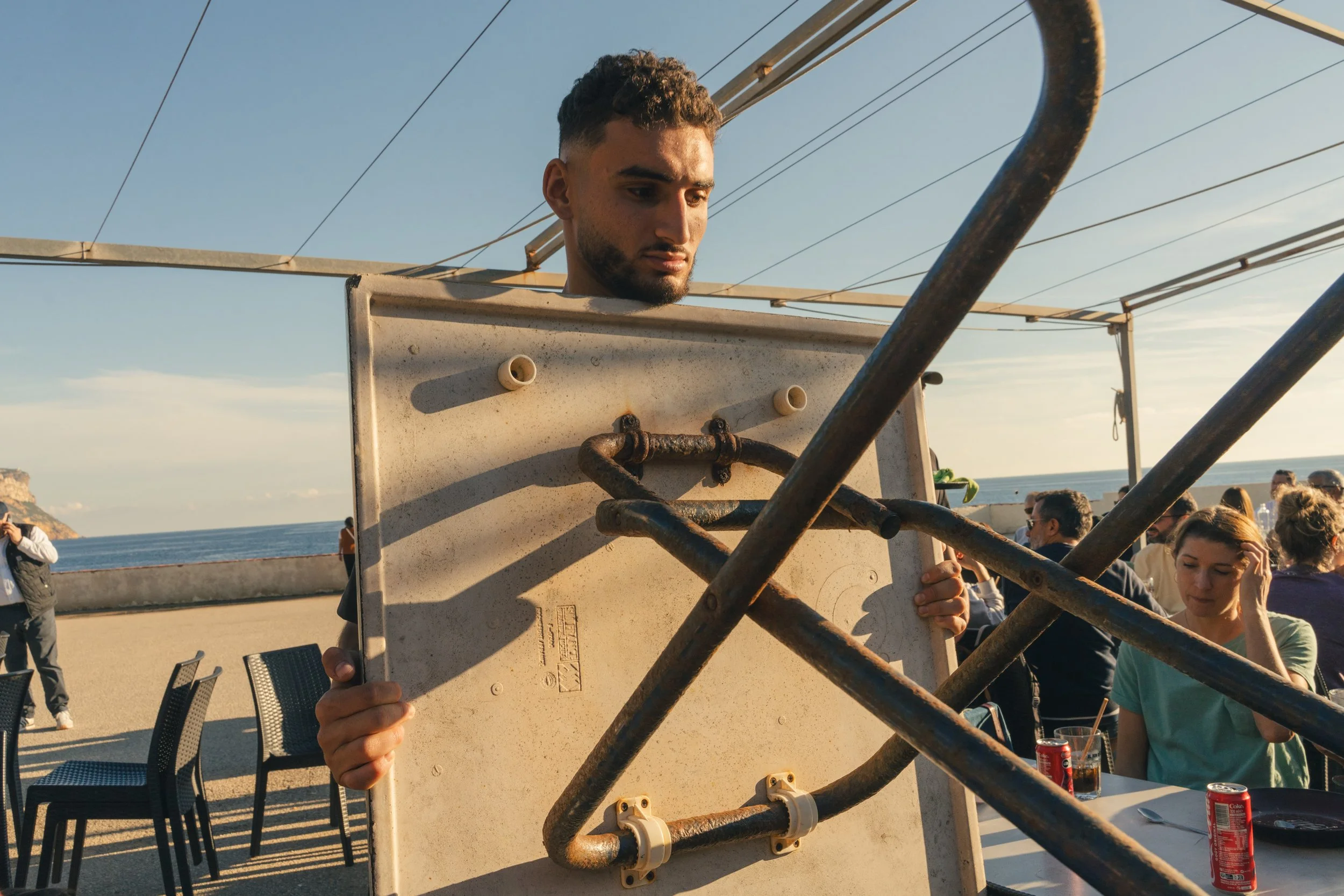 A man holding an old, rusty metal frame at an outdoor restaurant by the ocean, with other people seated and enjoying drinks in the background as the sun sets.