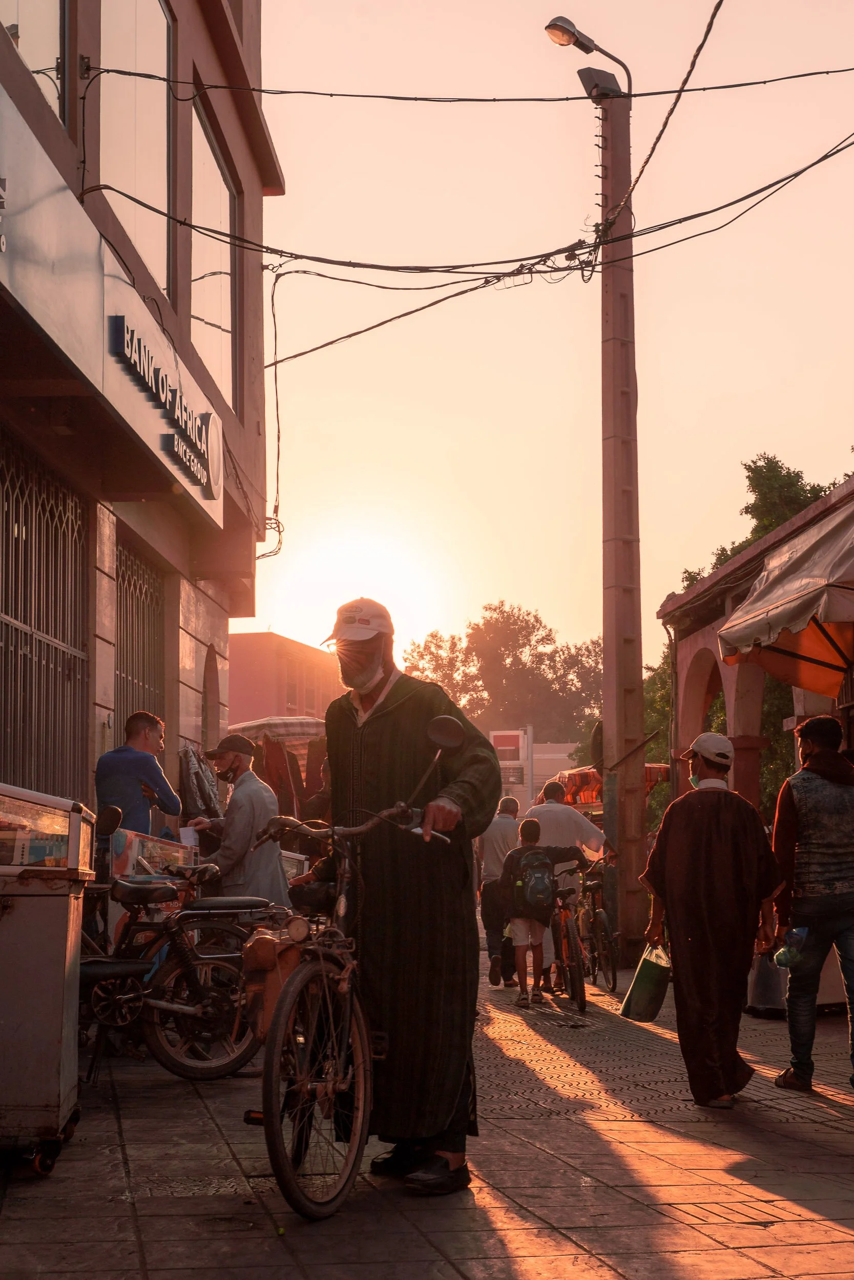 A busy street scene at sunset with people walking, some wearing masks, and a man in traditional attire with a bicycle in the foreground. There are buildings, street lamps, and power lines visible.