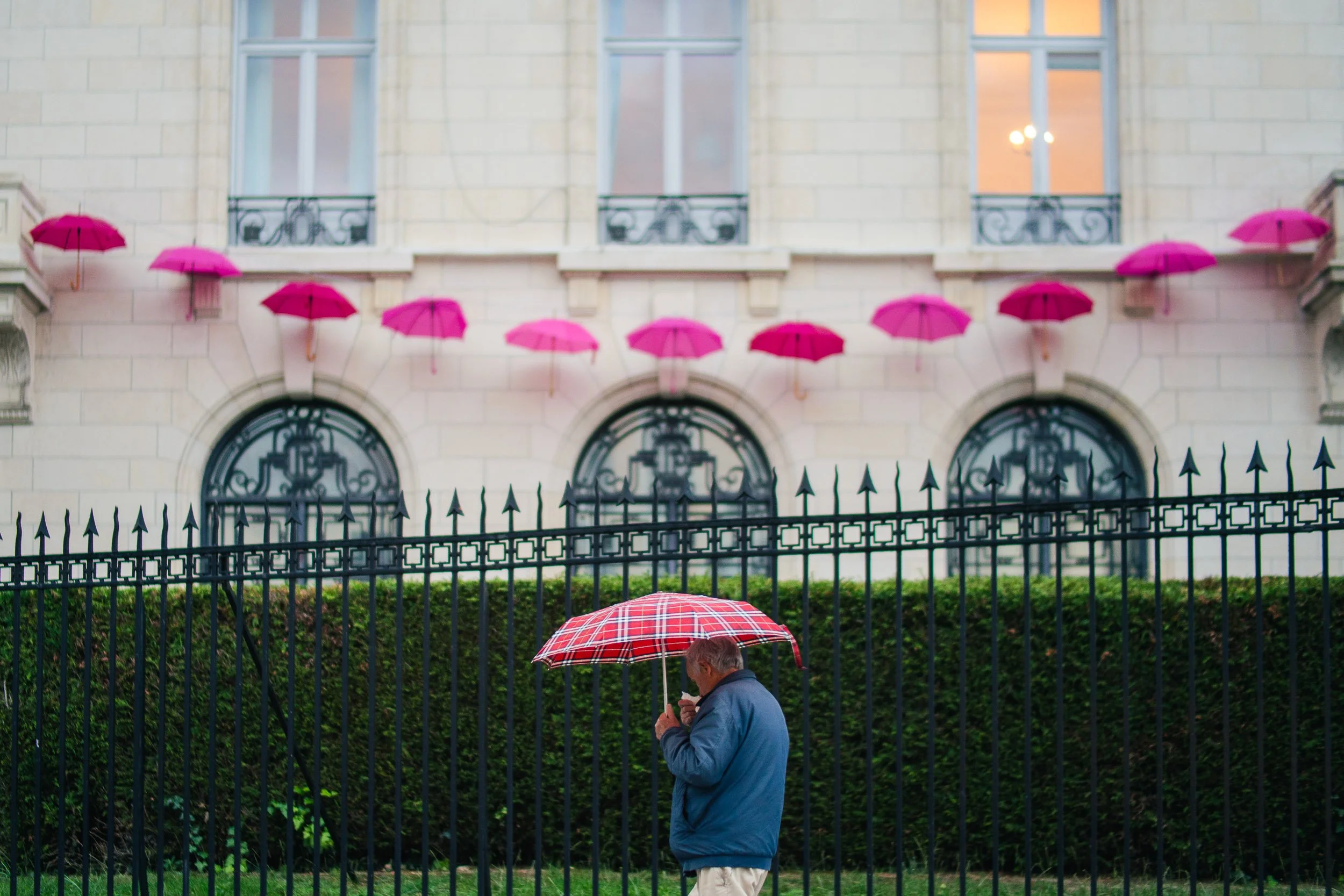 A man holding a red plaid umbrella standing behind a black wrought-iron fence, with the exterior of a large, ornate building in the background. The building has several pink umbrellas attached to its windowsills and is illuminated from inside.