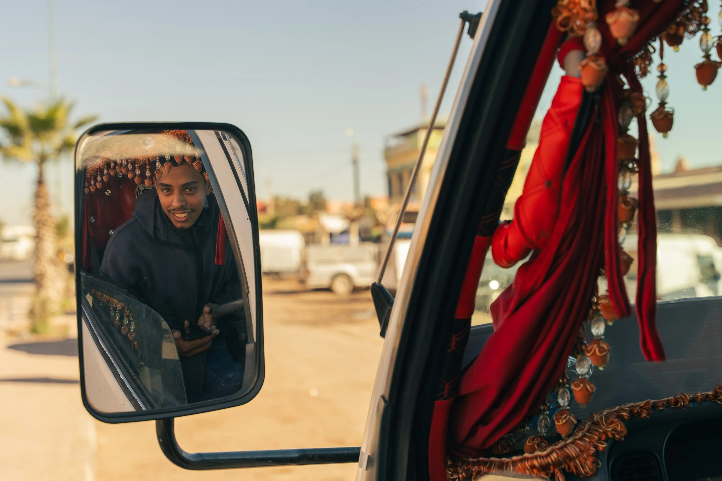 A young man looking into a vehicle's side mirror, smiling, with decorative red curtains hanging inside the vehicle.