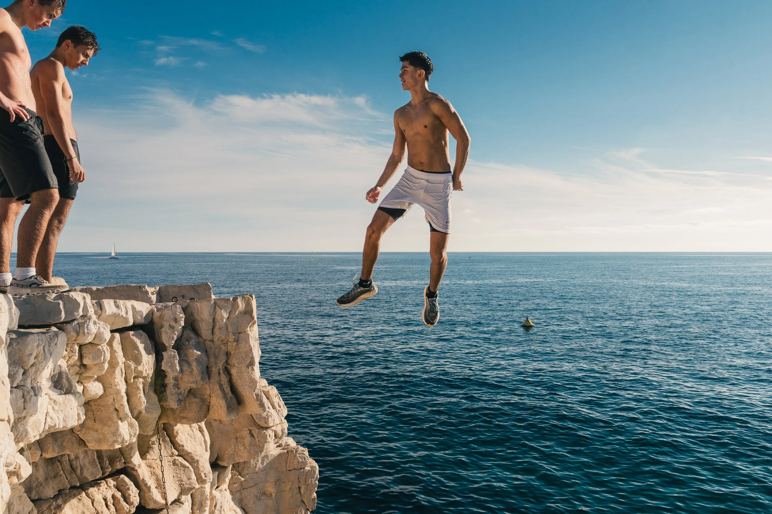 A young man jumps off a rocky cliff into the ocean while three other men watch from the edge on a sunny day.