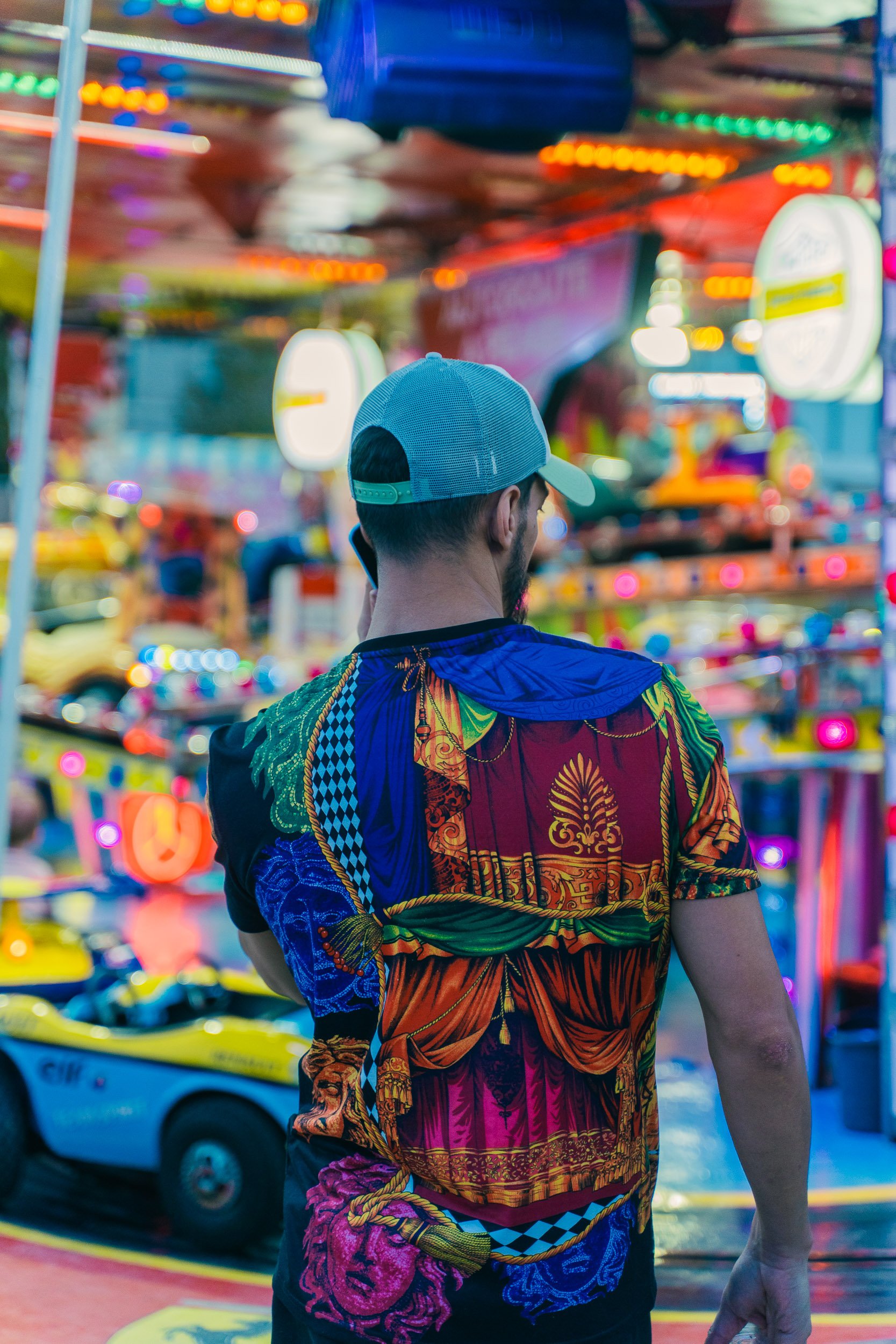 Back of a man in a colorful patterned shirt and a gray cap, standing in a brightly lit carnival or amusement park.