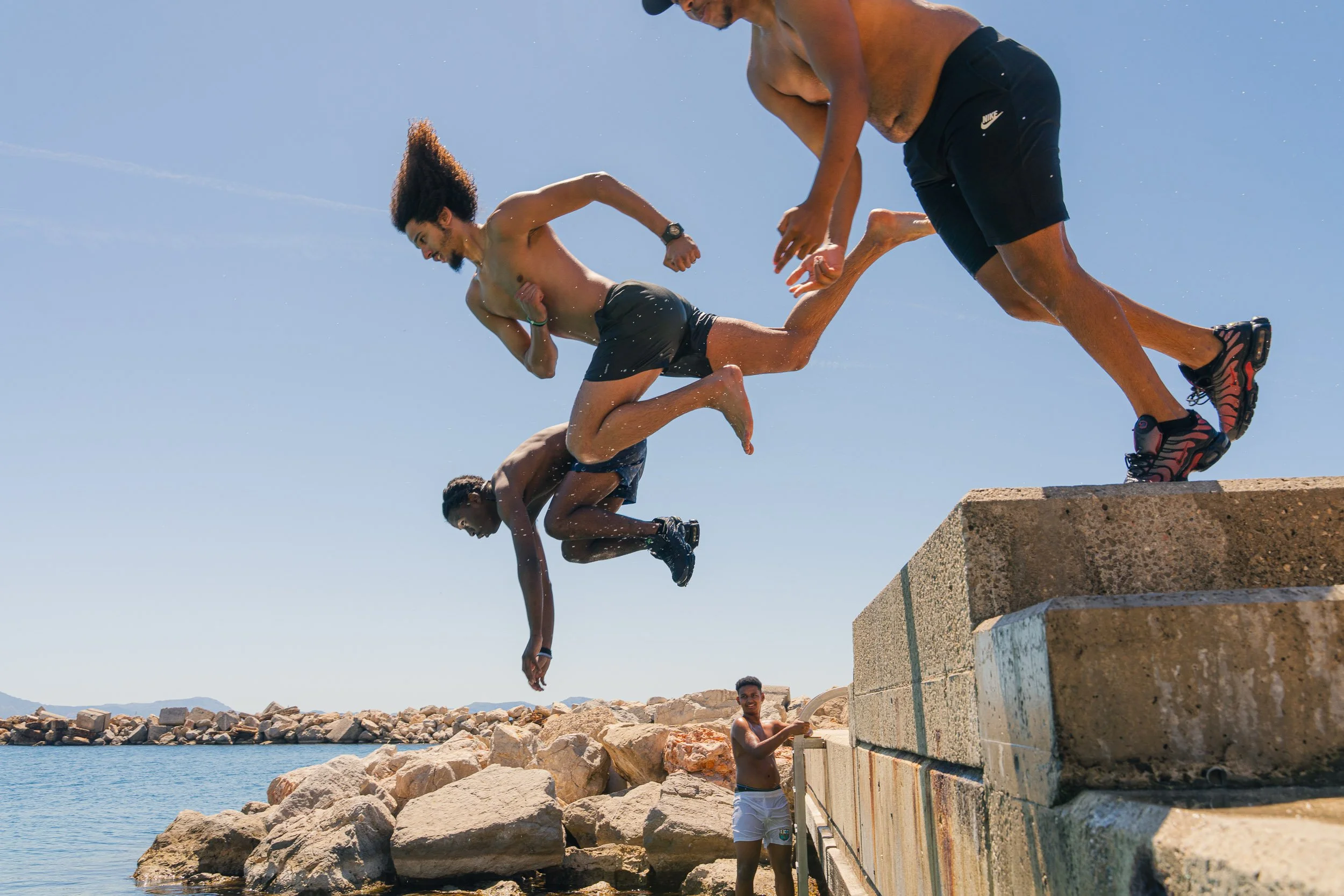 Group of young men in swim trunks jumping from a concrete dock into water at the beach on a sunny day.