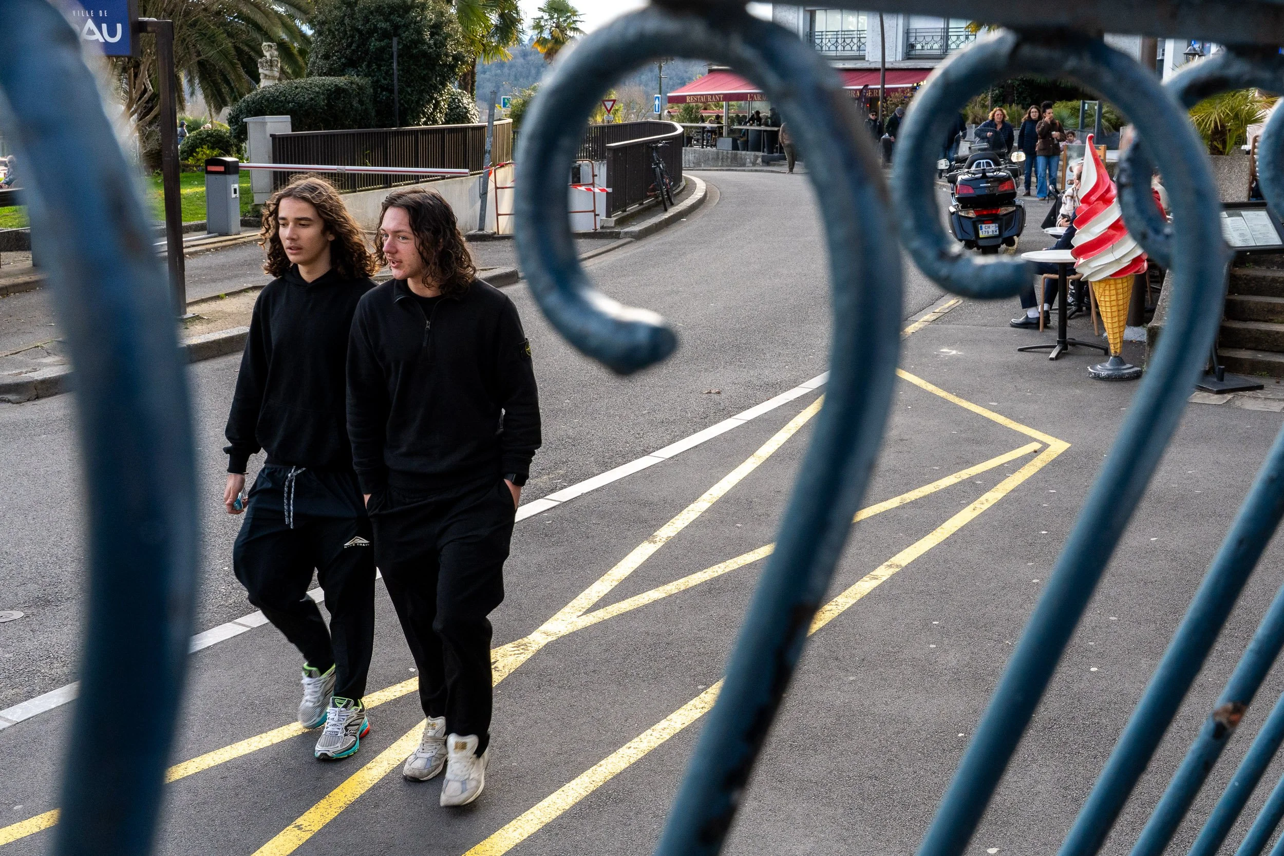 Two young people walking on a street near a cafe or ice cream stand, seen through a blue metal fence.