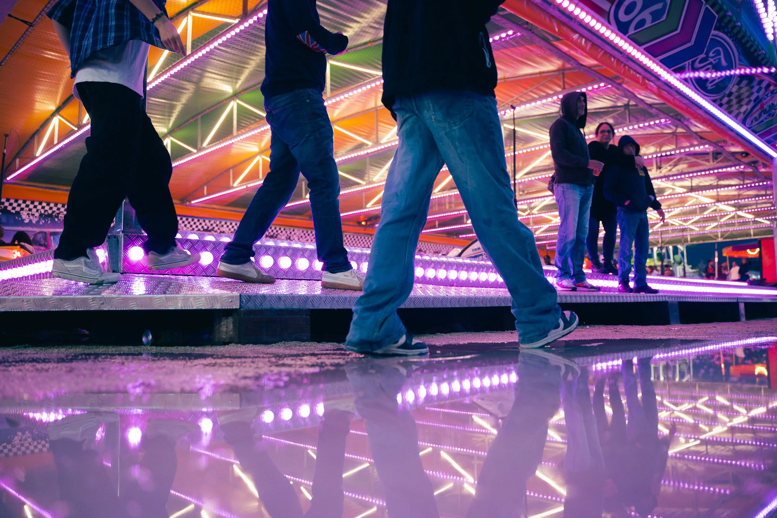 People walking in line on a spinning amusement park ride at night with bright neon purple and orange lights, reflected on the wet ground.