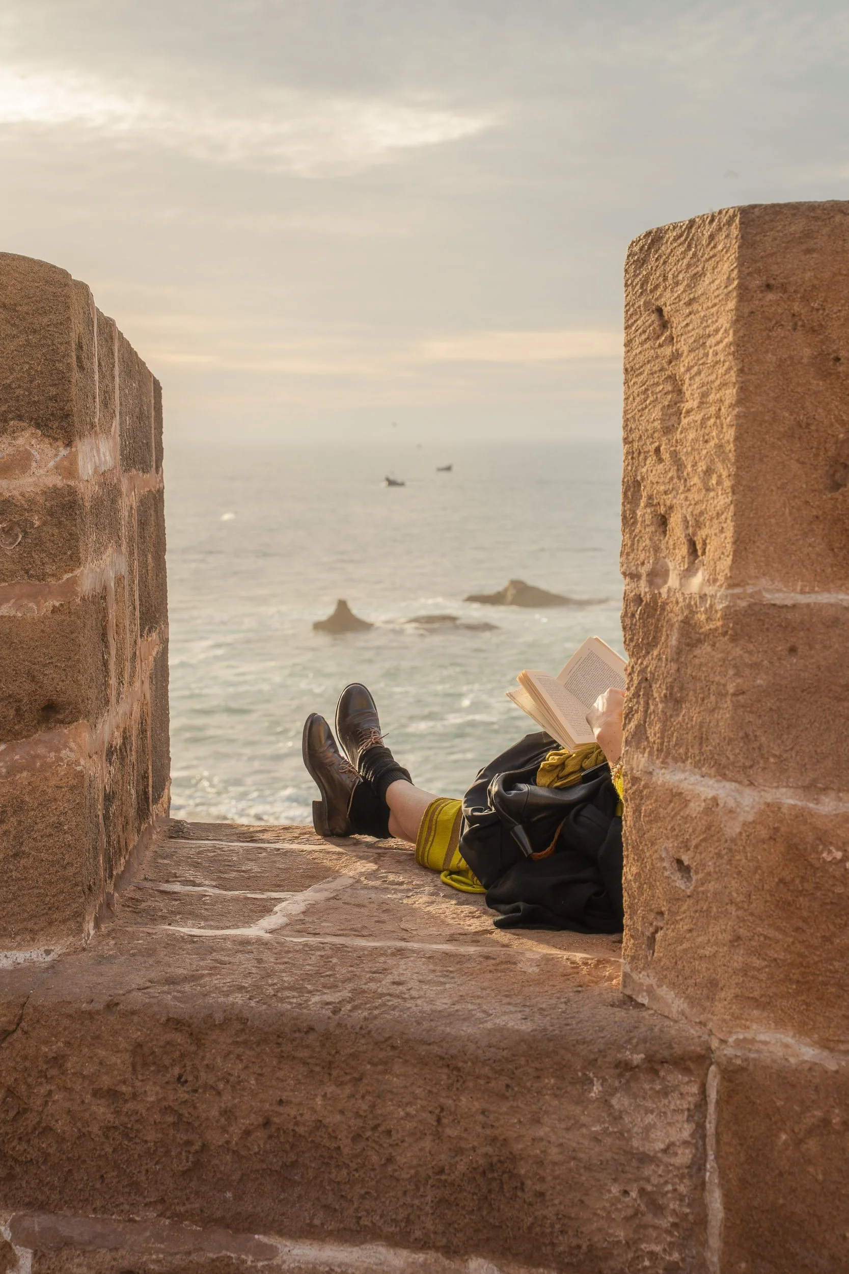 Person lying on a stone ledge by the sea, reading a book, with legs crossed and wearing black shoes, surrounded by brick walls, during sunset.