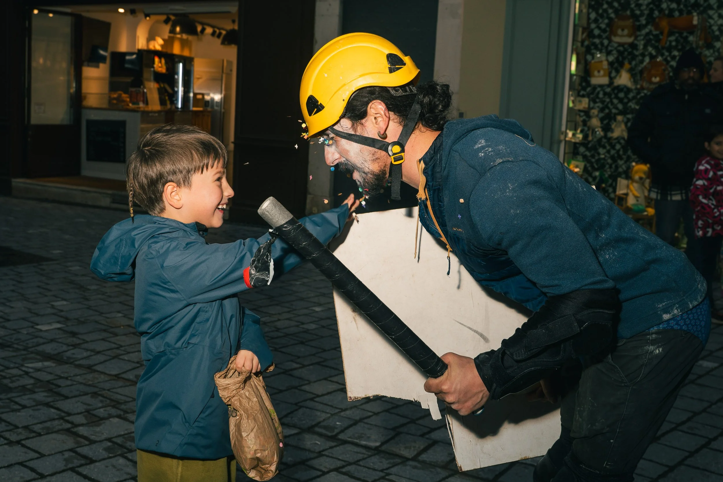 A young boy and a man wearing a yellow helmet and holding a large foam hammer, engage in a playful confrontation on a city sidewalk at night, with festive lights and shopfronts in the background.